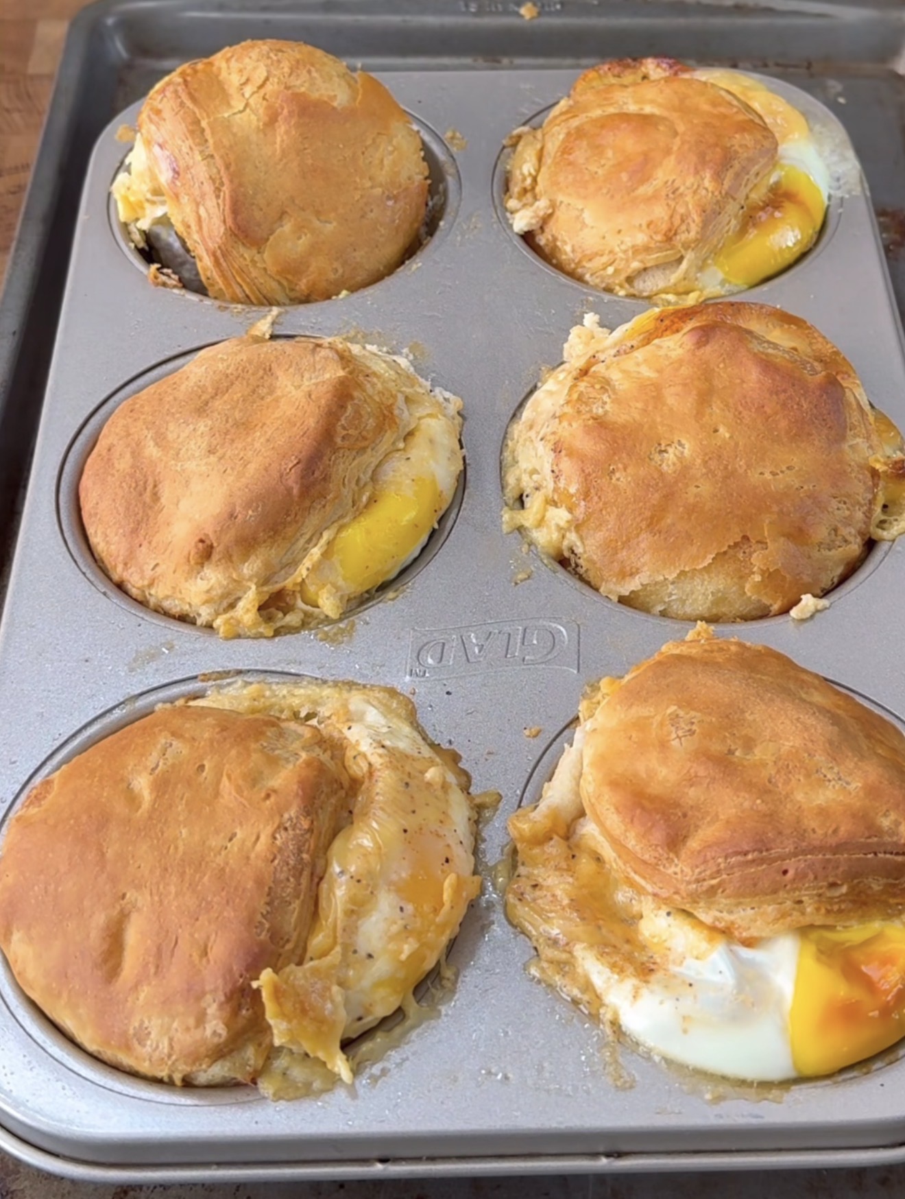 A muffin tin filled with six baked biscuit cups, each containing eggs and possibly cheese, with golden-brown biscuit tops and some egg spilling out from the edges.