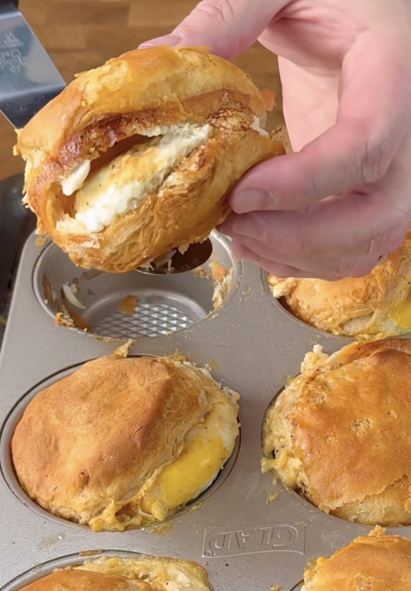 A hand holds a baked biscuit filled with egg and cheese above a muffin tin, which contains more golden, fluffy biscuit breakfast cups.