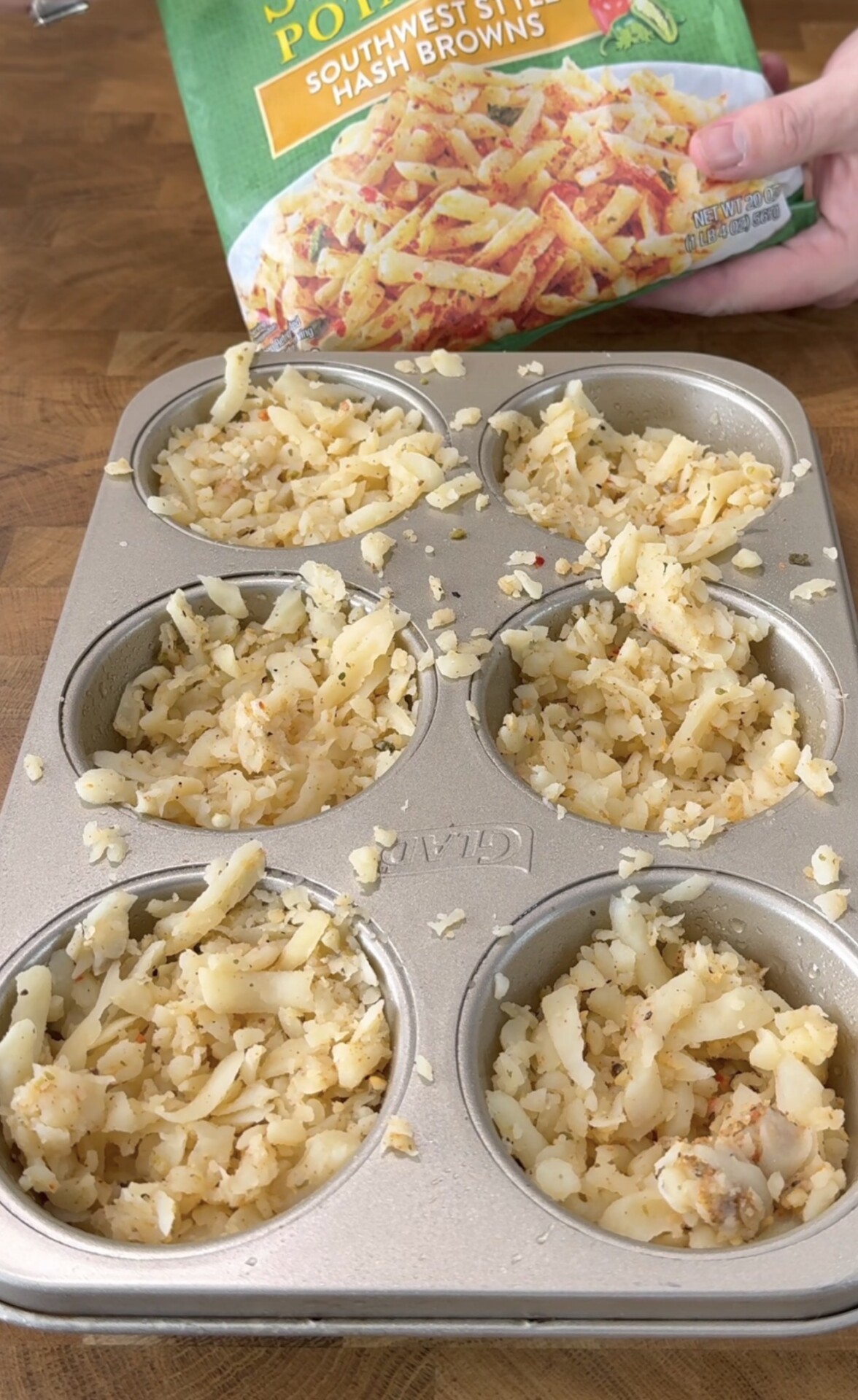 A muffin tin filled with shredded hash browns. A hand holds a bag labeled “Southwest Style Hash Browns” above the tin on a wooden surface.