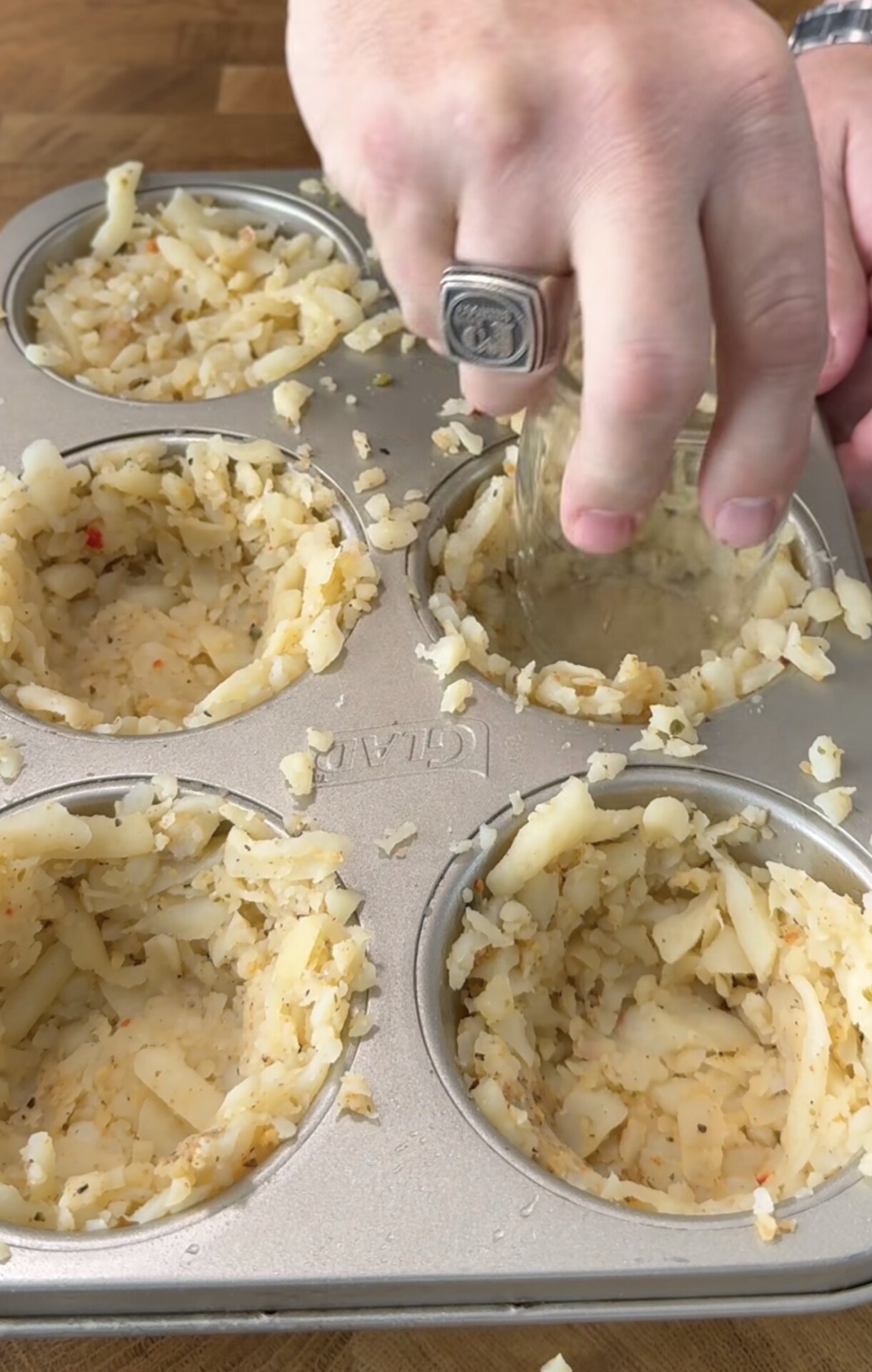 A hand presses shredded potatoes into muffin tin cups using a small glass to form nests, preparing them for baking. The muffin tin sits on a wooden surface.