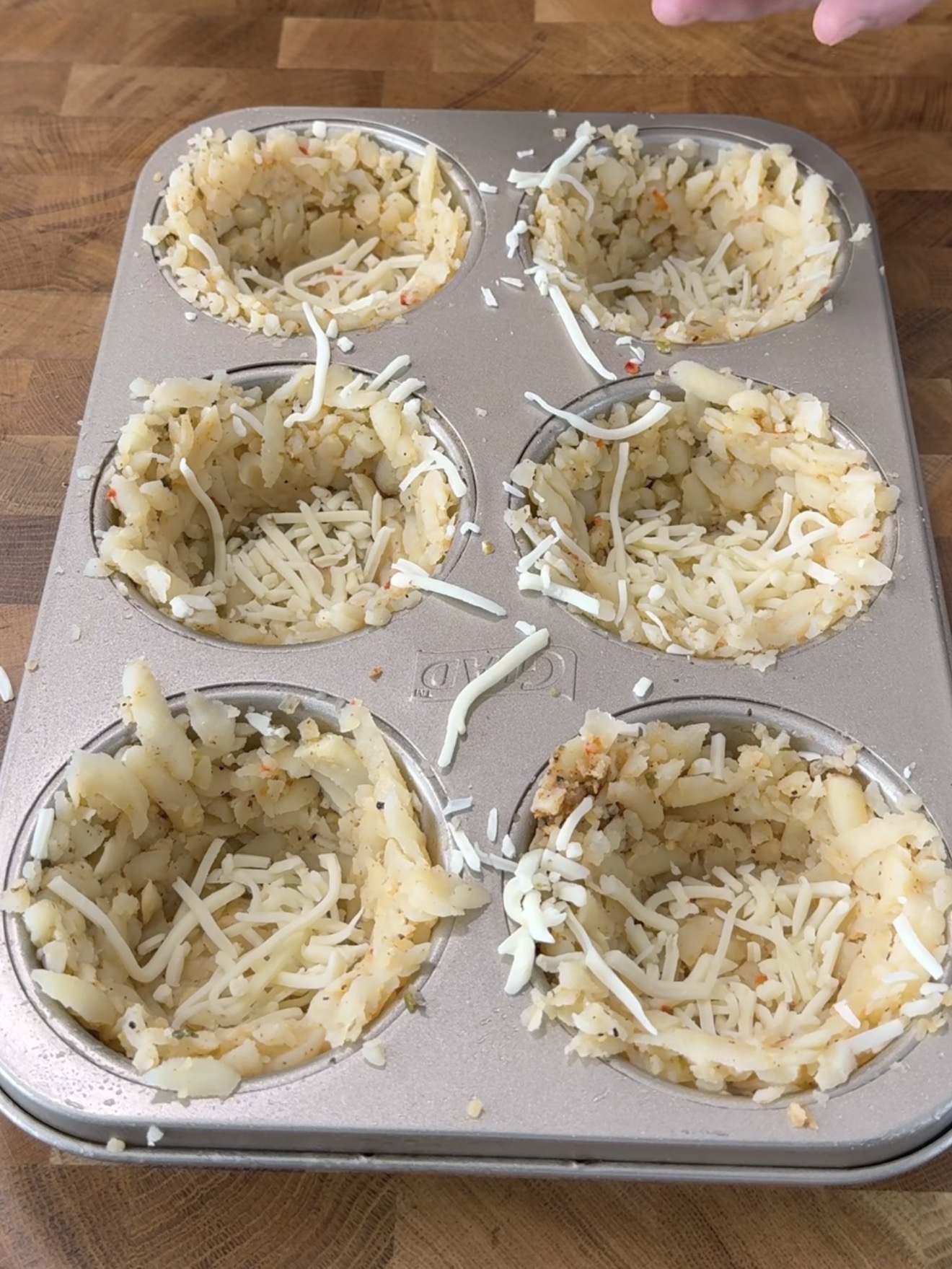 A muffin pan with six cups lined with shredded potatoes and sprinkled with grated cheese, forming nest-like shapes, sits on a wooden surface.