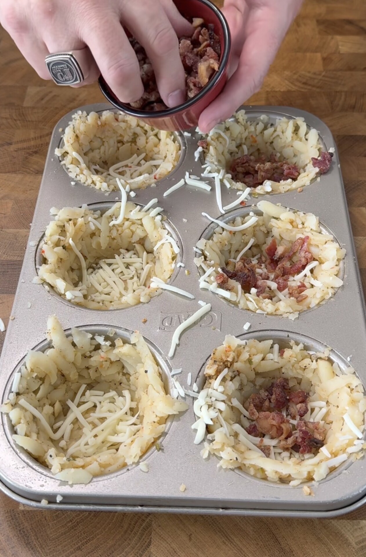 A hand sprinkles bacon bits into shredded potato cups topped with cheese in a muffin tin on a wooden surface.