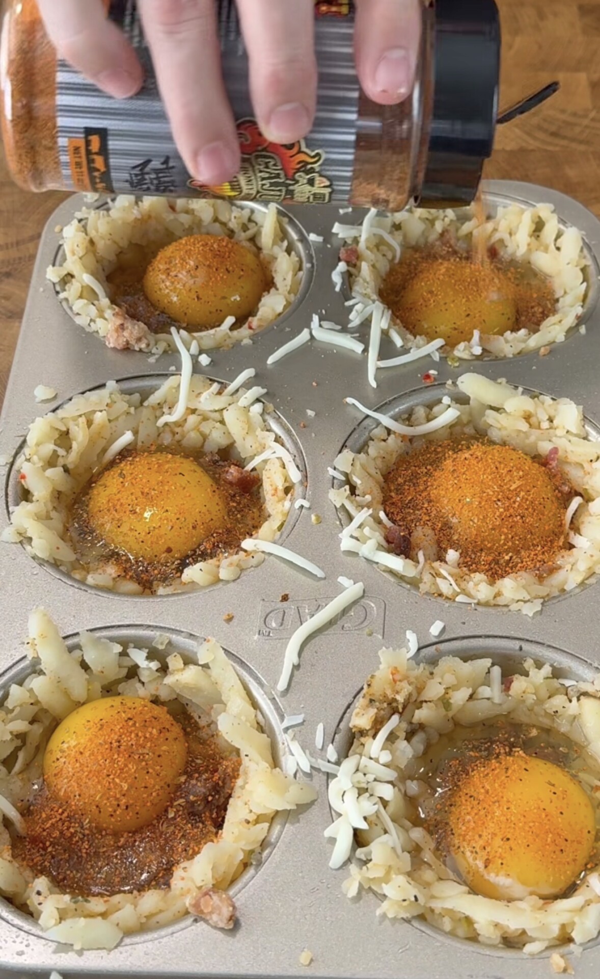 A hand sprinkles seasoning over raw eggs placed in hash brown nests inside a muffin tin, preparing a breakfast dish on a wooden surface.