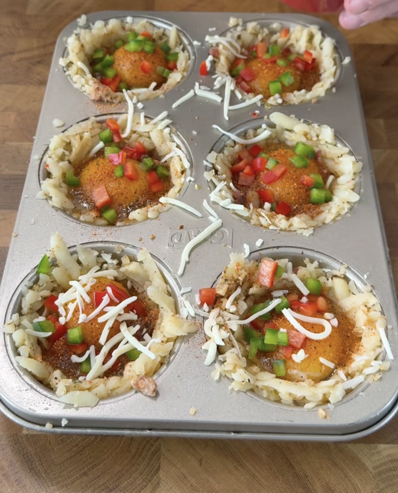 A muffin tin filled with six nests of shredded potatoes, each holding an egg topped with shredded cheese and diced red and green bell peppers, sits on a wooden surface.