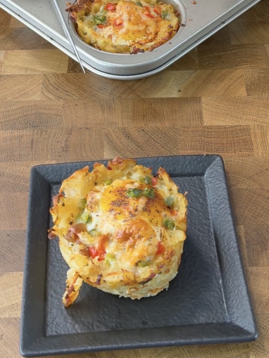 A baked egg muffin filled with cheese, red and green peppers, and herbs sits on a black square plate, with a muffin tin containing more muffins in the background on a wooden surface.