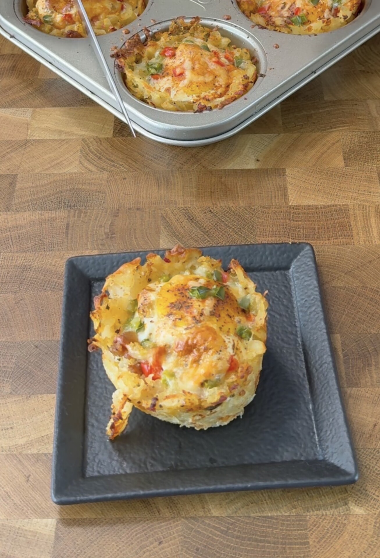A baked noodle muffin topped with melted cheese and chopped vegetables sits on a square black plate, with more muffins in a metal muffin pan in the background on a wooden surface.
