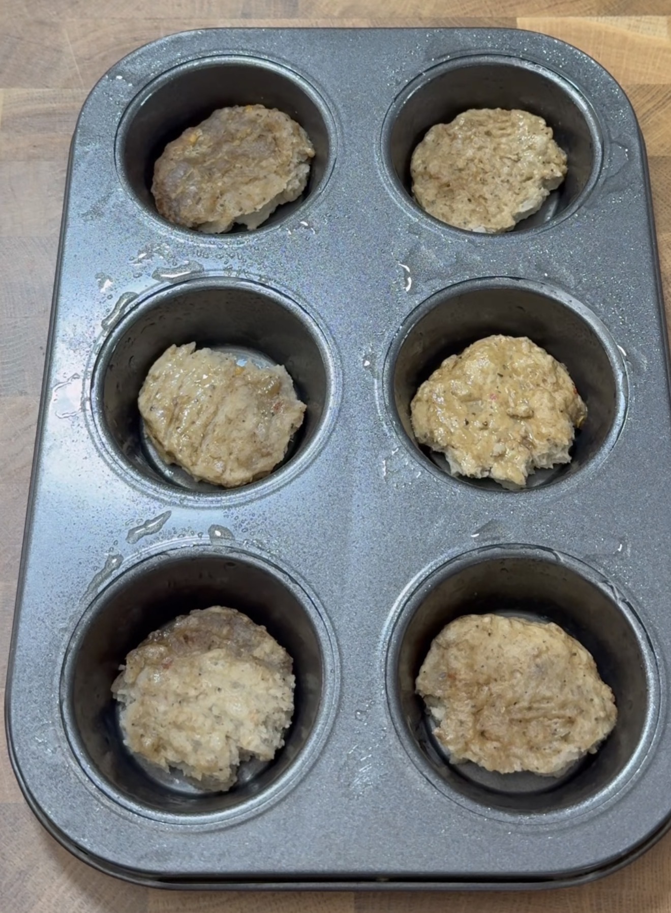 A muffin tin with six round, browned patties or meat mixtures, each placed in a separate cup, sits on a wooden surface.