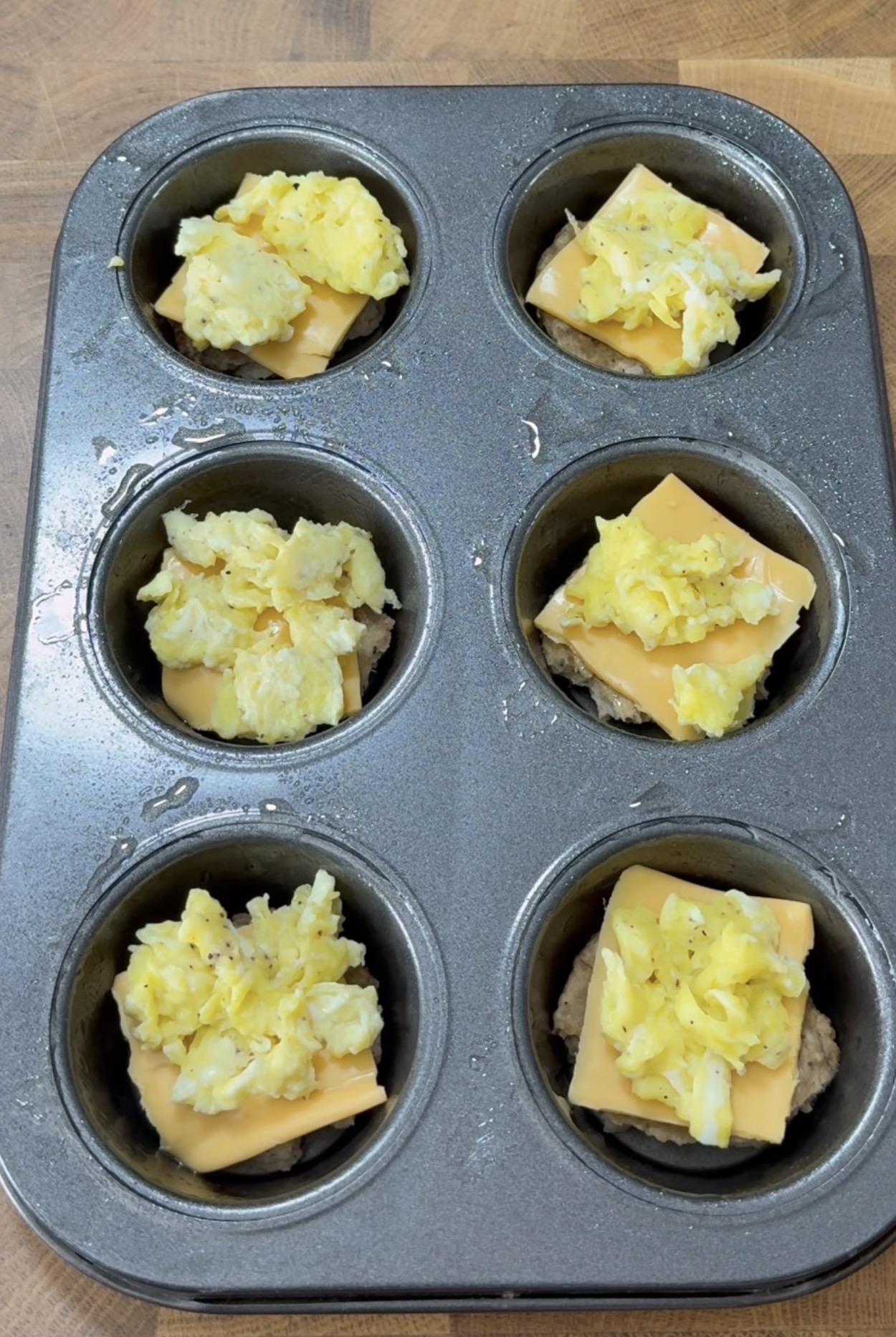 A muffin tin with six cups, each holding a layer of sausage patty, a slice of cheddar cheese, and a scoop of scrambled eggs, sits on a wooden surface, ready for baking.