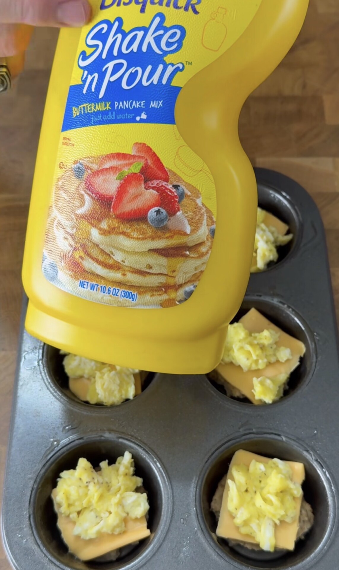 A hand holds a Bisquick Shake n Pour Buttermilk Pancake Mix bottle above a muffin tin filled with scrambled eggs, cheese slices, and sausage patties on a wooden surface.