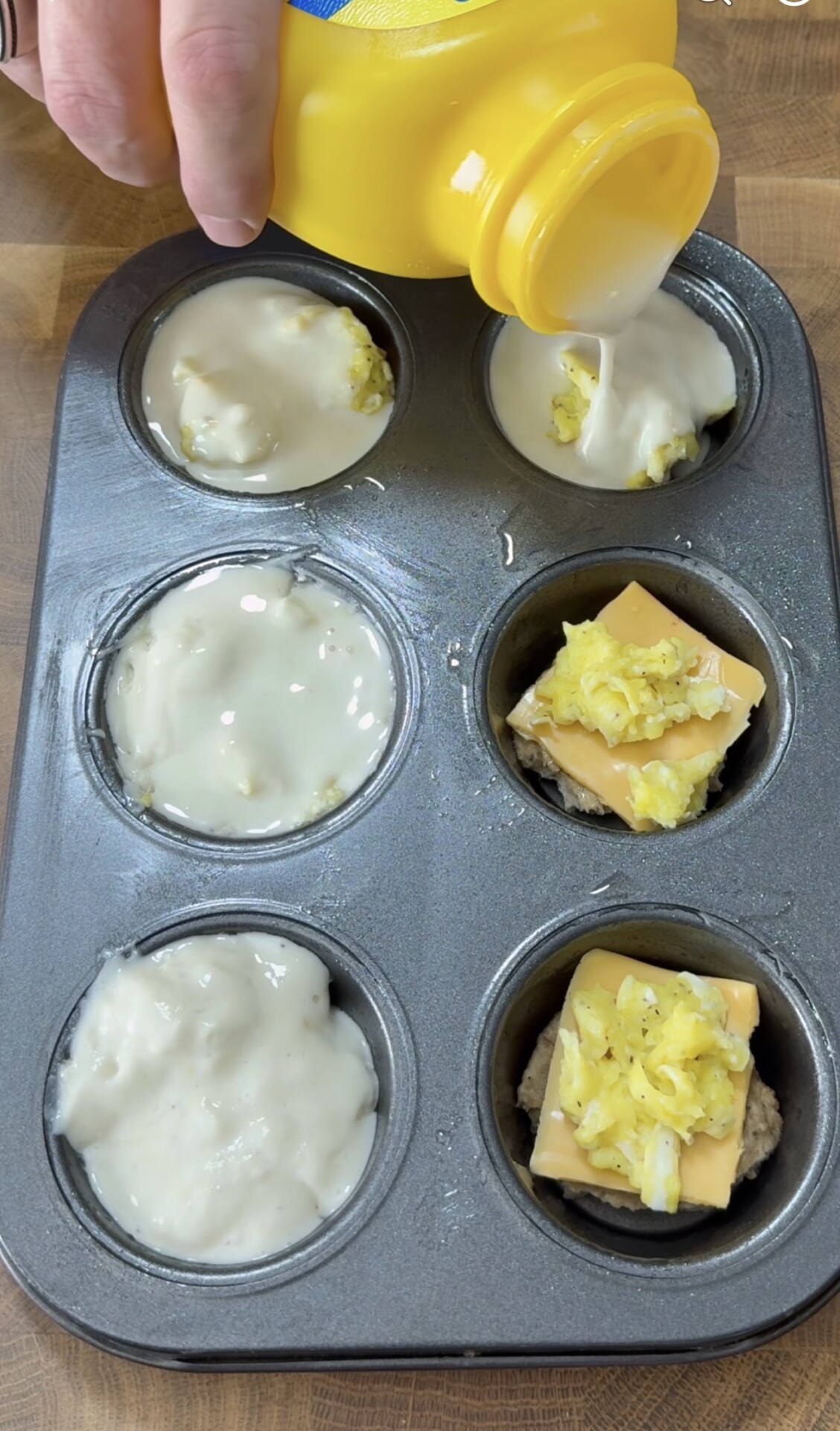 A hand pours batter from a yellow container into a metal muffin tin. Some cups contain batter, while others have a slice of cheese and cooked scrambled eggs on a sausage patty. The tin is on a wooden surface.