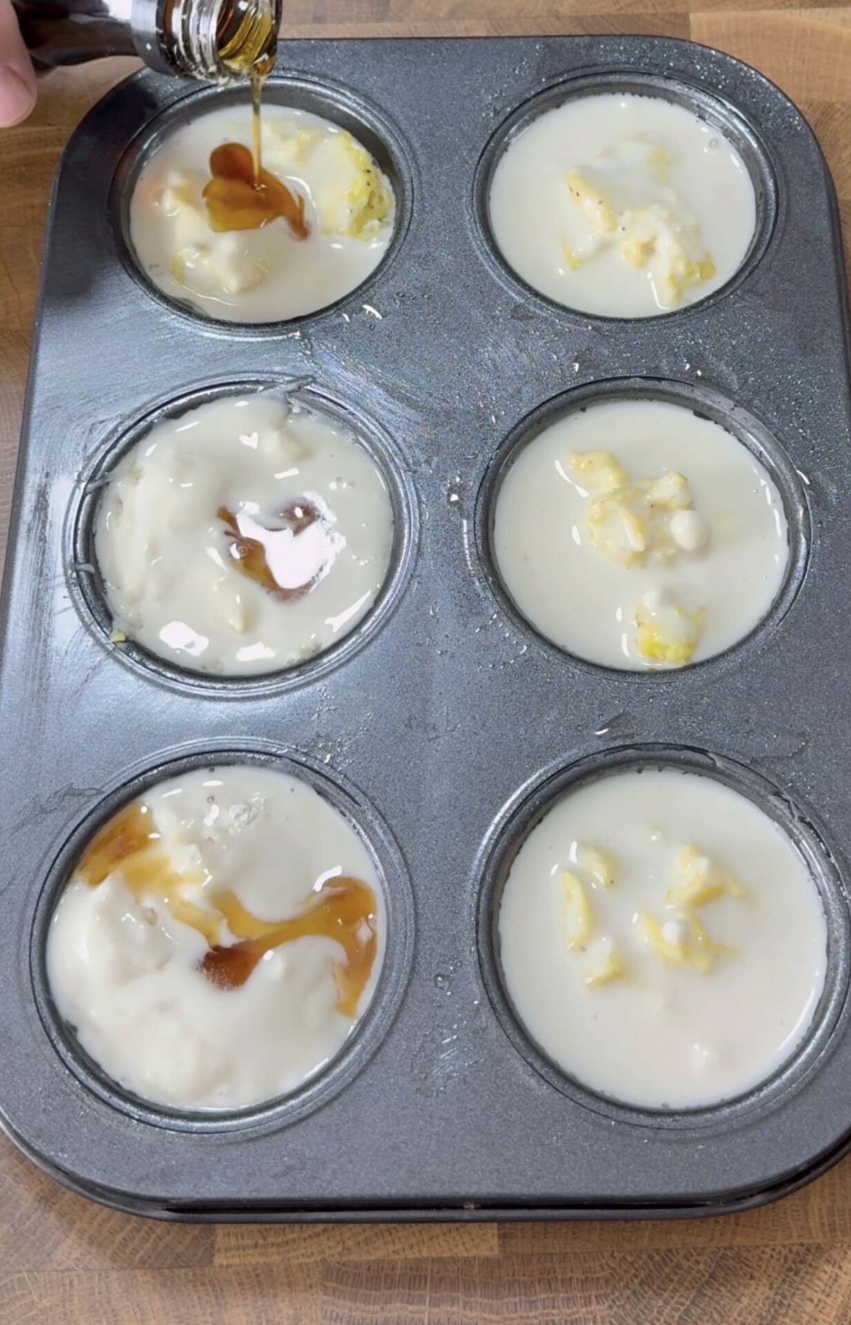 A muffin tin with six cups is filled with light batter, some containing chunks. A hand pours a brown liquid, possibly vanilla, into one cup. The tin sits on a wooden surface.