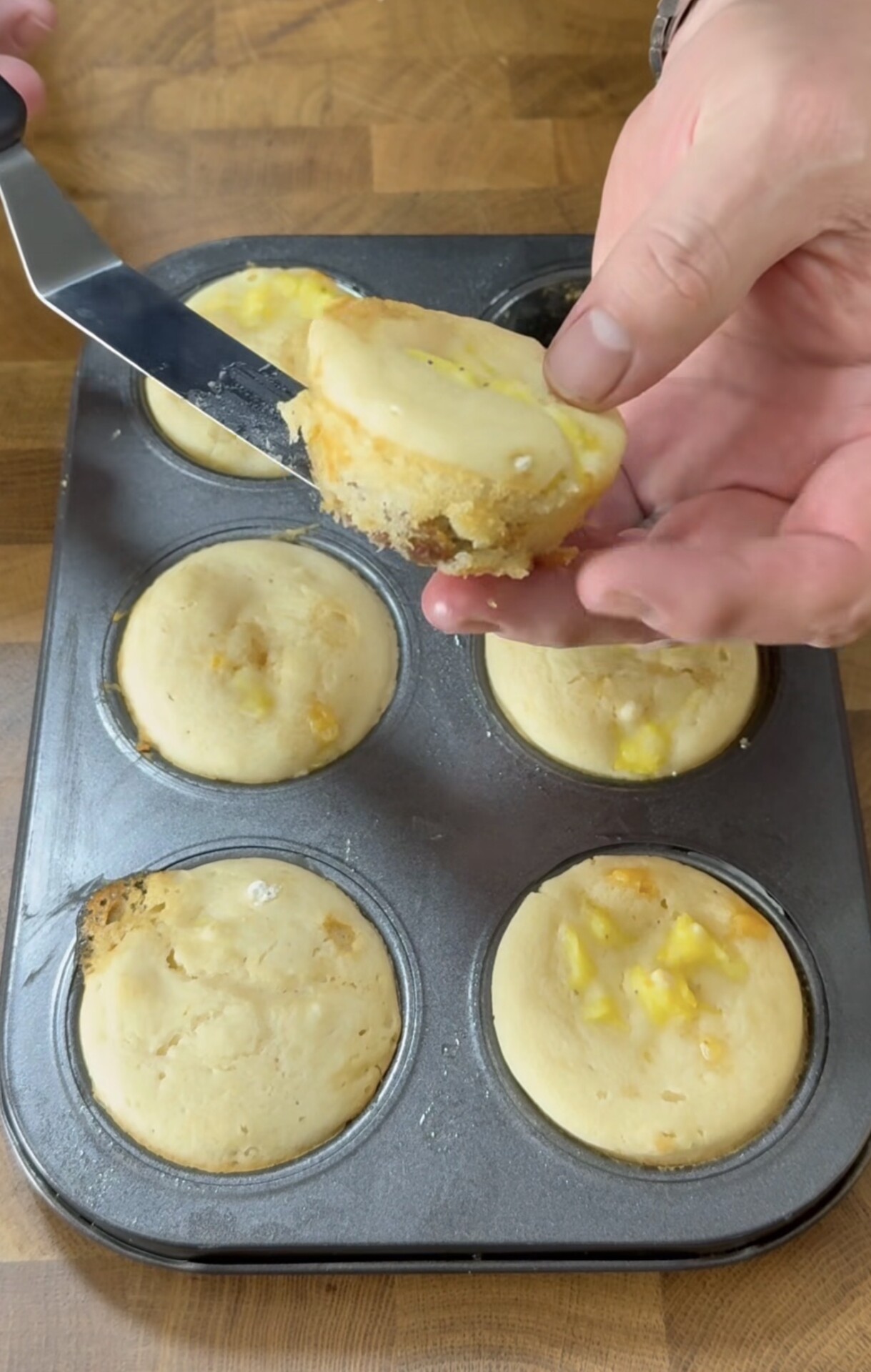A hand lifts a freshly baked muffin out of a muffin tin using a spatula. The muffins are light golden and have visible chunks of fruit inside. The muffin tin rests on a wooden surface.