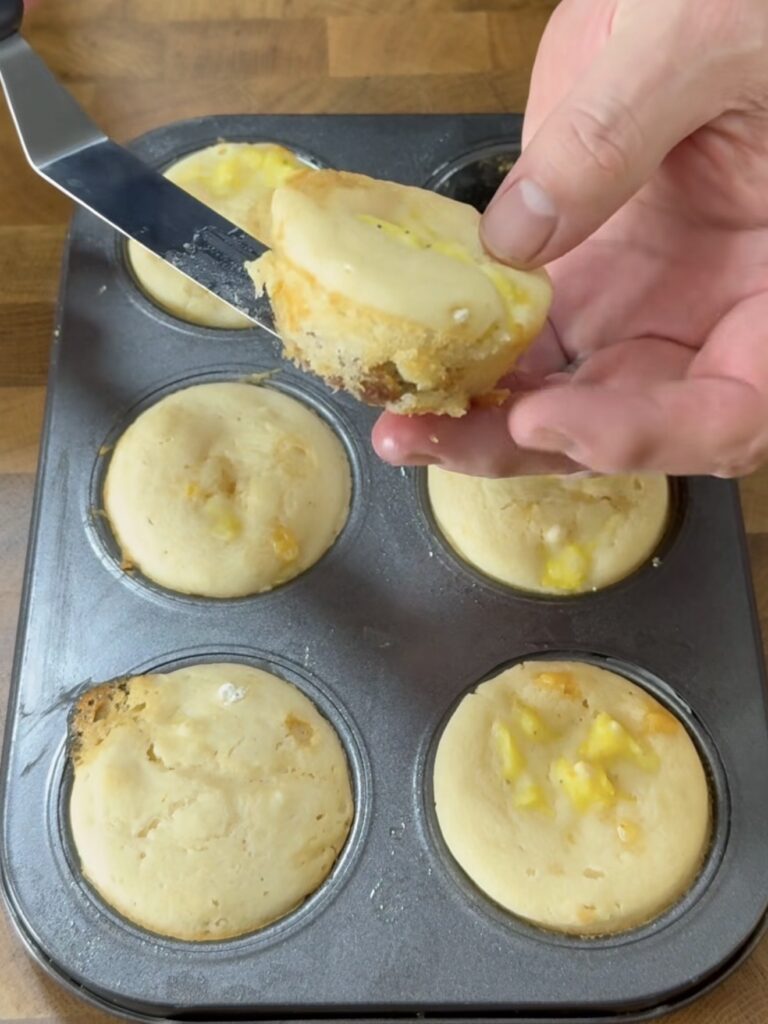 A hand uses a spatula to lift a muffin from a metal muffin tin containing six partially baked muffins with visible yellow fruit pieces on a wooden surface.
