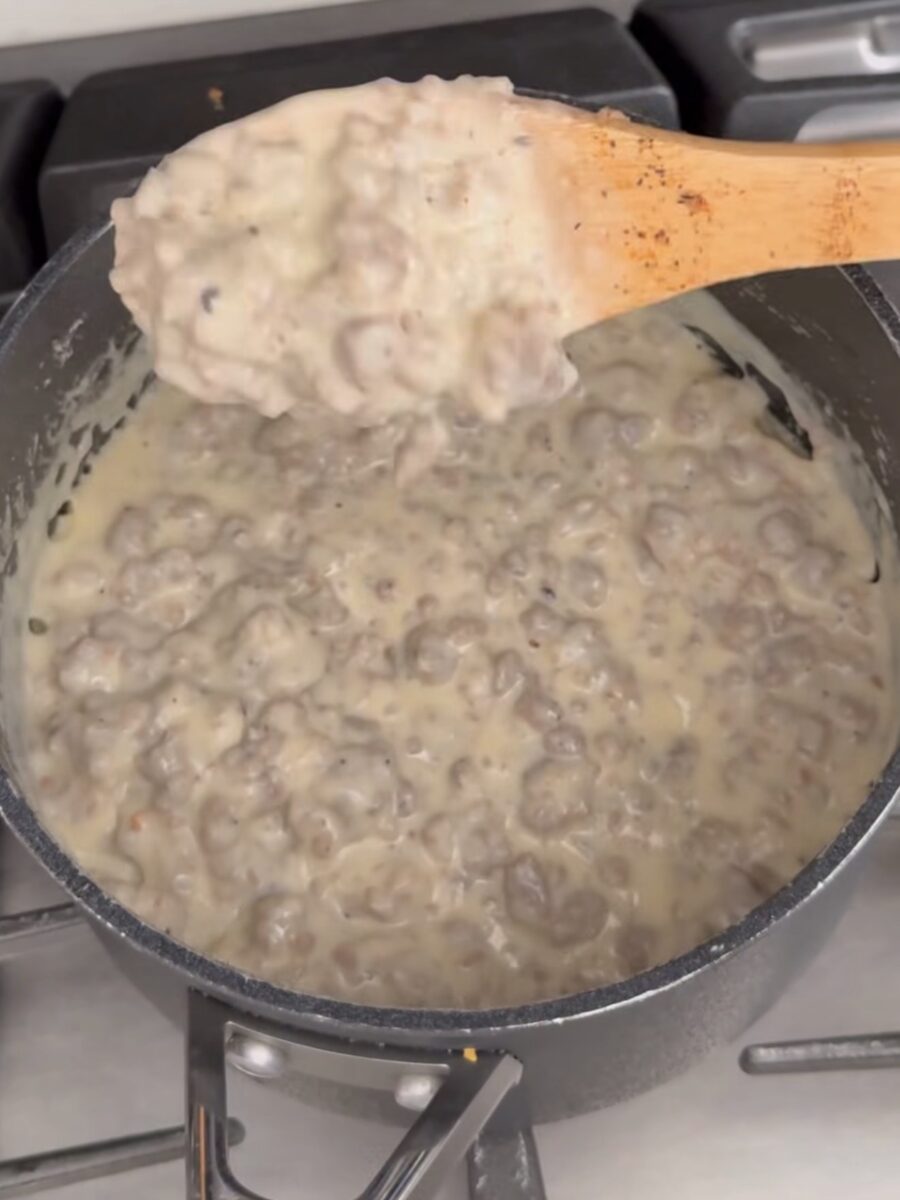 A pot of creamy sausage gravy is being stirred with a wooden spoon over a stovetop. The thick, white gravy is filled with chunks of cooked sausage.