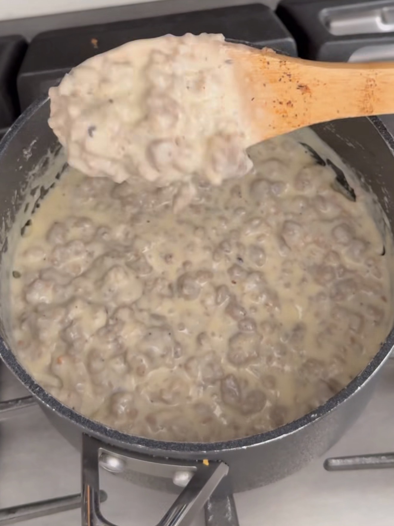 A pot of creamy sausage gravy is being stirred with a wooden spoon over a stovetop. The thick, white gravy is filled with chunks of cooked sausage.