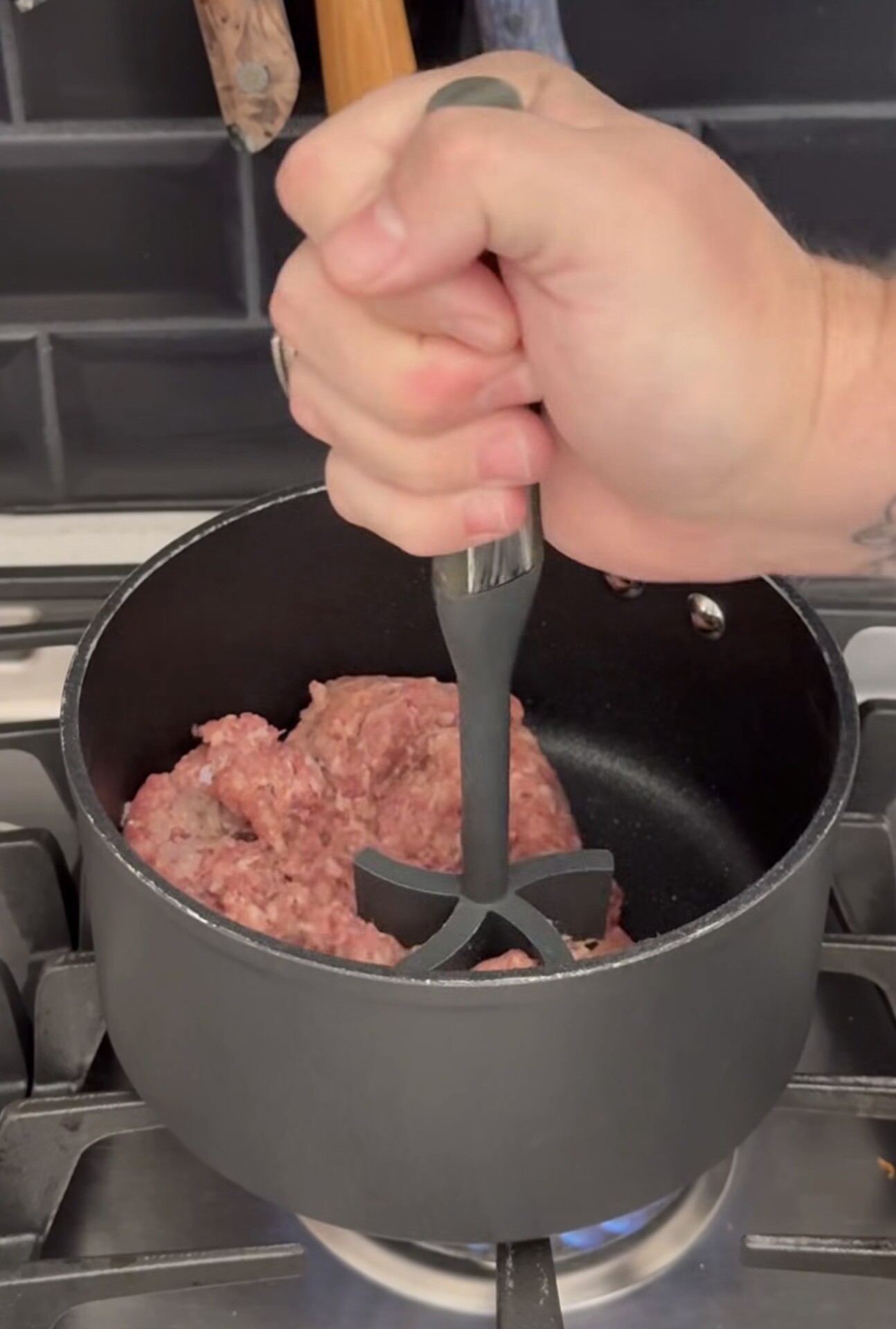 A person uses a meat chopper to break up raw ground meat in a black pot on a gas stove.