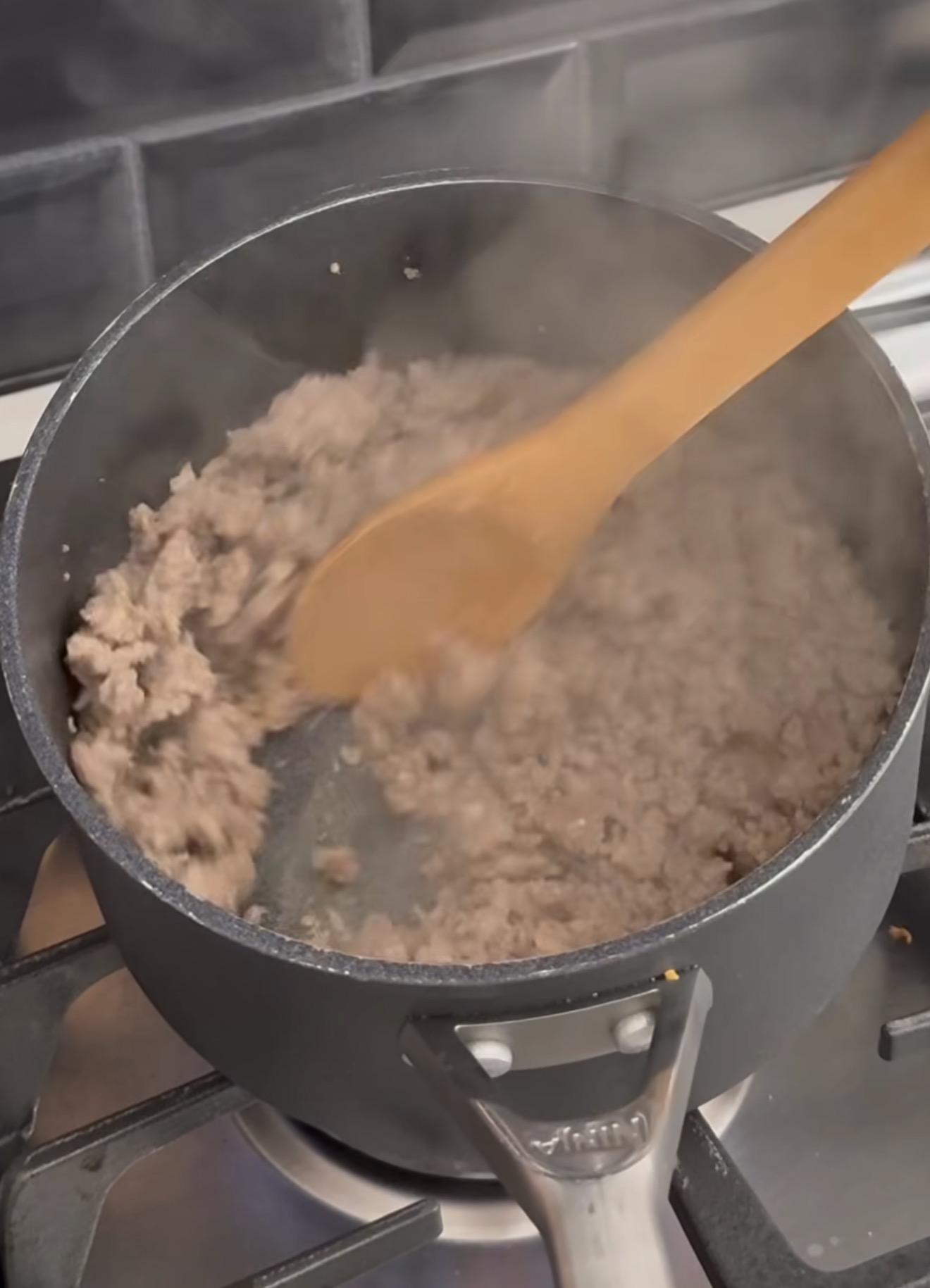 Ground meat is being browned in a black pot on a stove, stirred with a wooden spoon. Steam rises from the pot, and black tile backsplash is visible in the background.