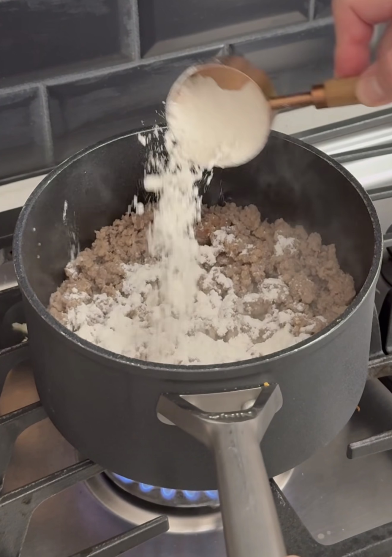 A hand sprinkles flour from a spoon over browned ground meat cooking in a black pot on a gas stove. Steam rises from the ingredients as they cook.