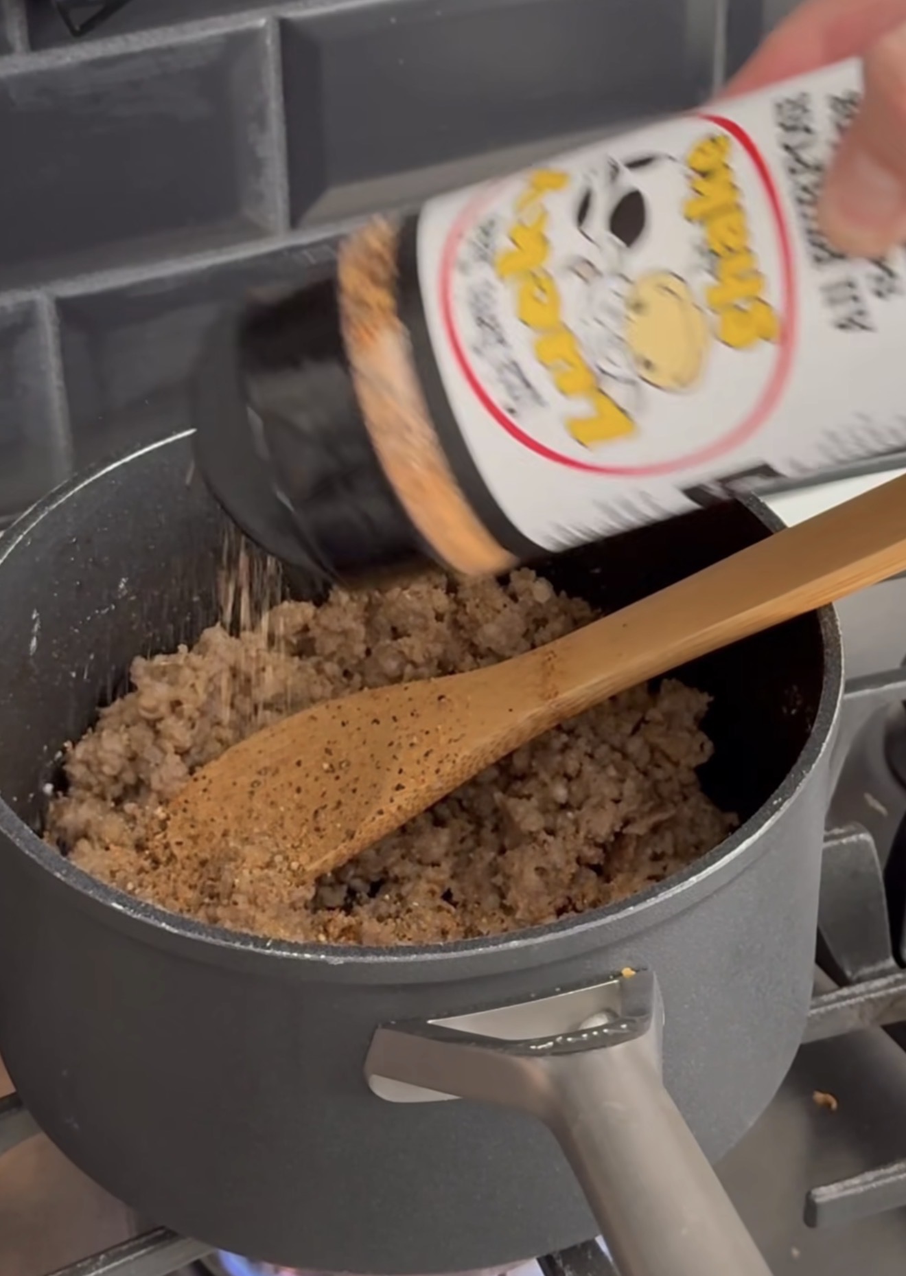 A hand sprinkles seasoning from a container labeled Lucky Shake onto cooked ground meat in a pot, stirring with a wooden spoon on a stove.
