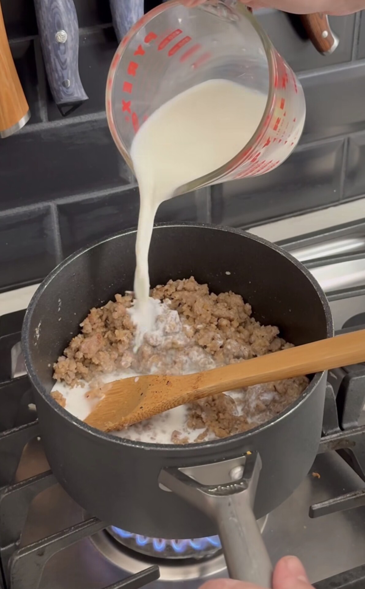 A person pours milk from a measuring cup into a pot of cooked ground meat on a stove, stirring with a wooden spoon.