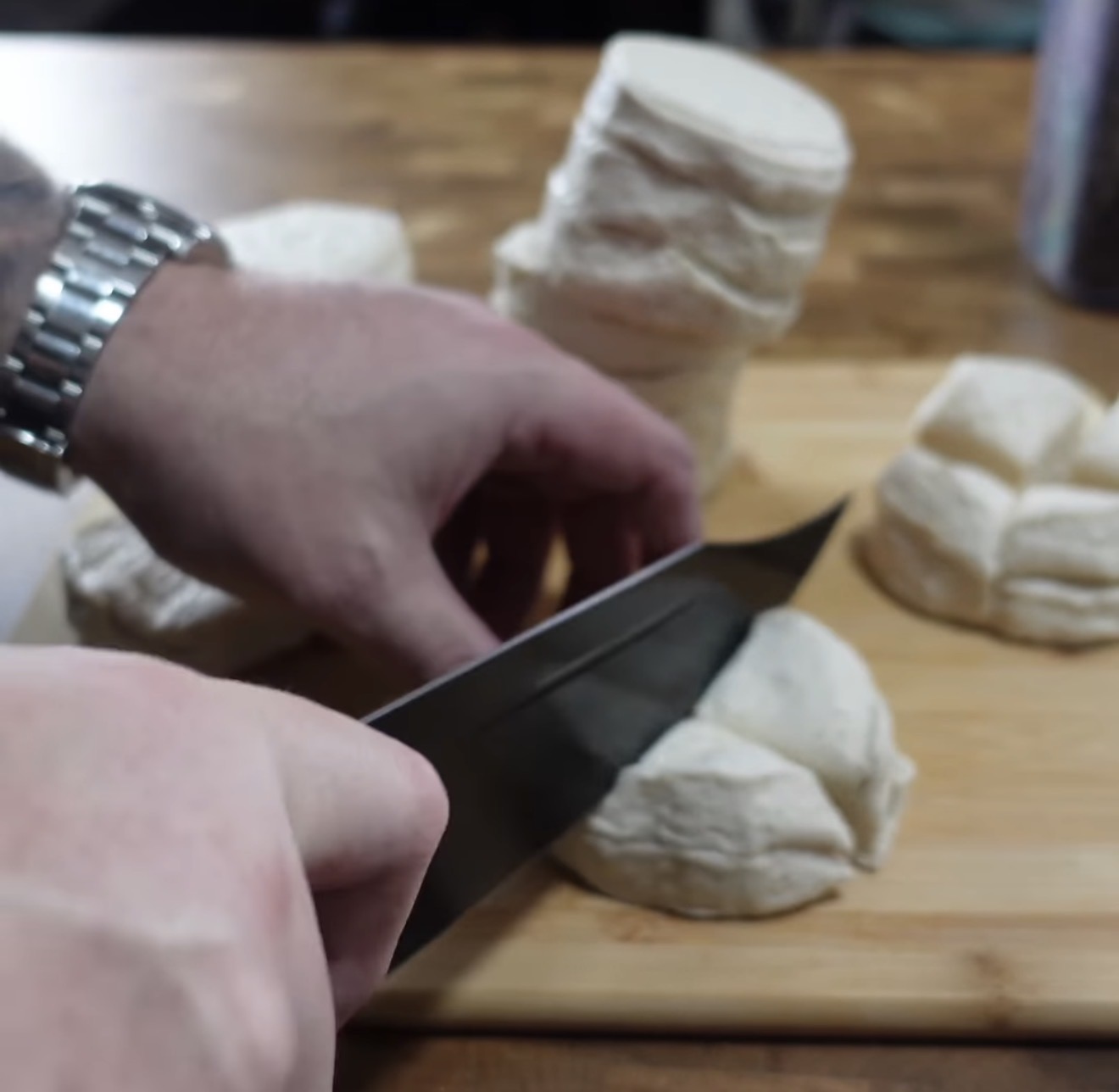 A person slices raw biscuit dough rounds into quarters on a wooden cutting board, with several stacks of dough in the background.