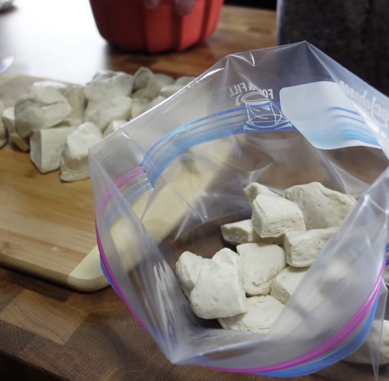 Chunks of raw biscuit dough are being placed from a wooden cutting board into a resealable plastic bag on a kitchen counter.