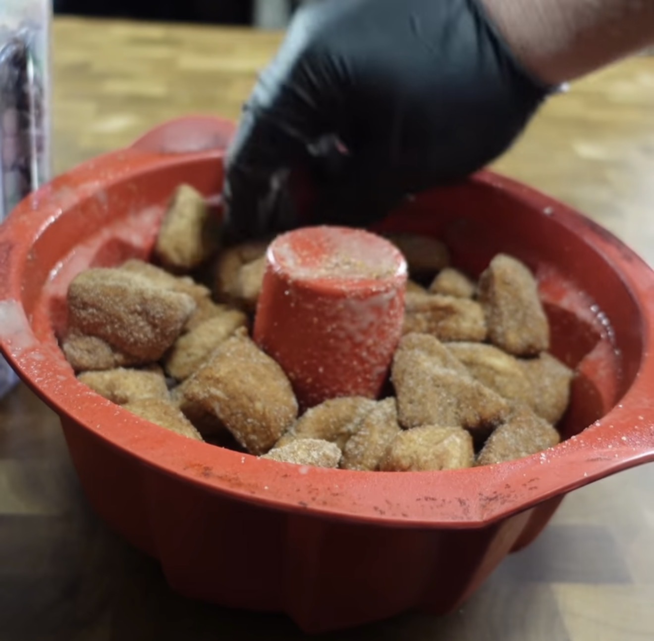 A gloved hand arranges sugar-coated dough pieces in a red Bundt pan on a wooden surface.