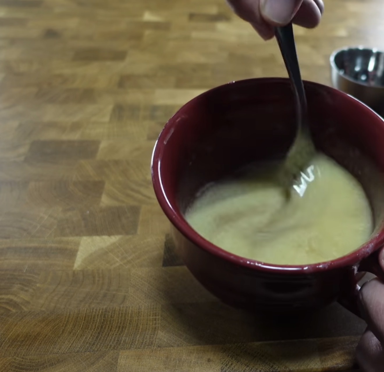 A hand stirs a light-colored mixture in a red bowl with a fork on a wooden countertop; a small metal bowl sits in the background.