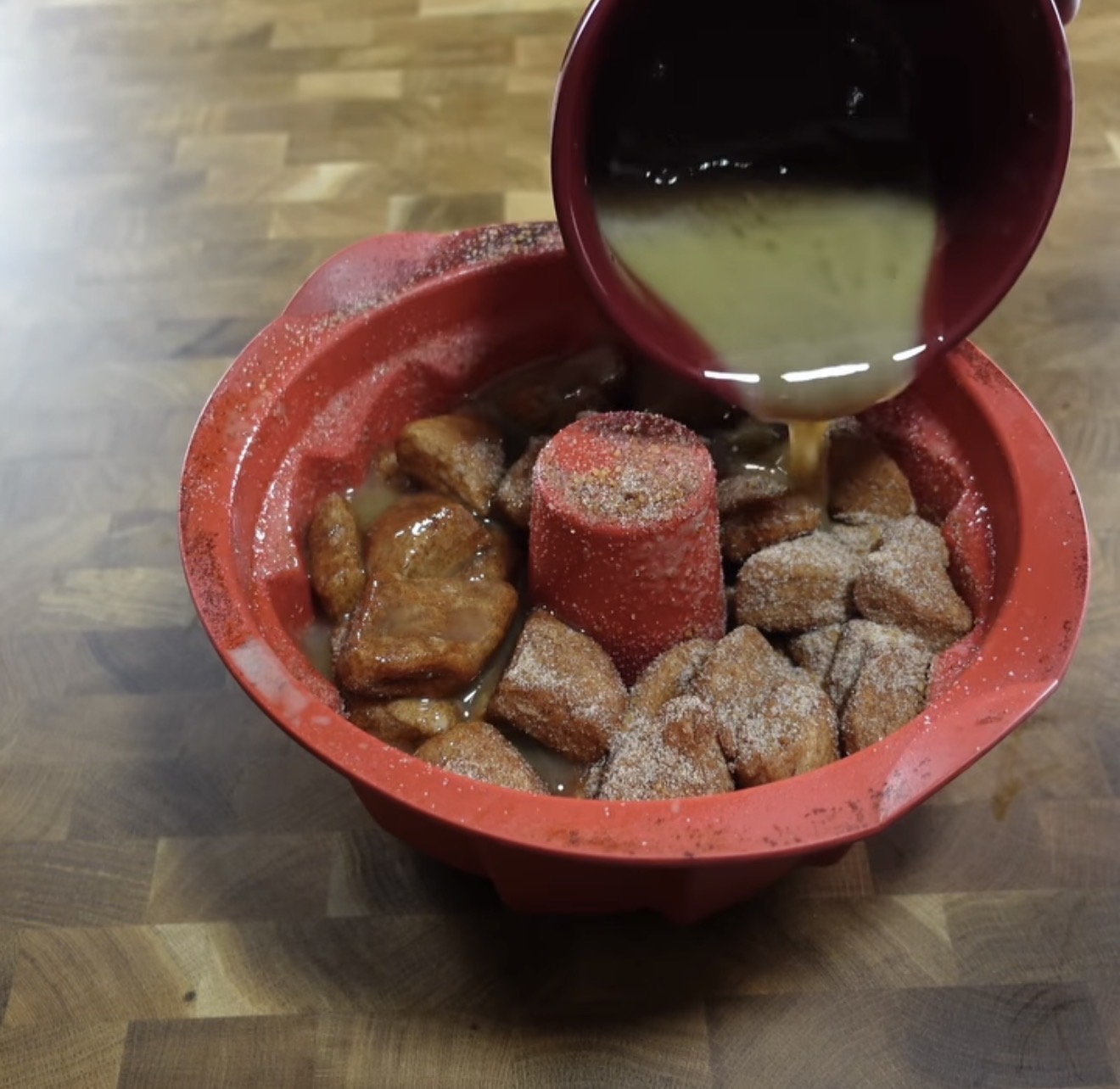 A red bundt pan filled with dough pieces coated in cinnamon sugar, with a light brown sauce being poured over them from a maroon bowl, all on a wooden surface.