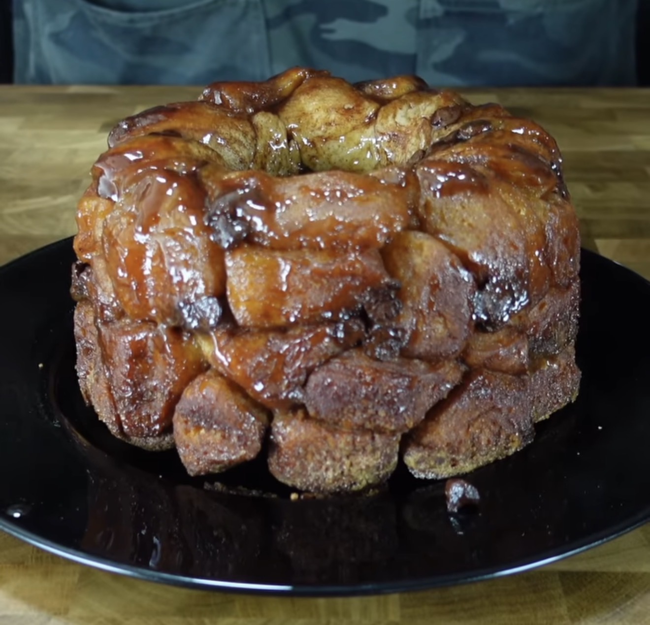 A sticky, caramel-coated monkey bread sits on a black plate atop a wooden surface, with a background of someone wearing a dark shirt.