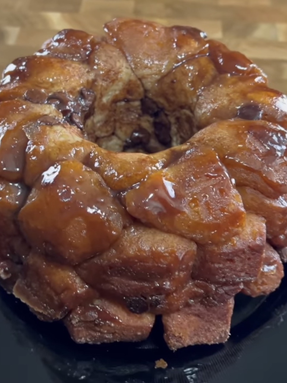 A close-up of a round, pull-apart monkey bread covered in a shiny caramel glaze, displayed on a dark plate with a wooden surface in the background.