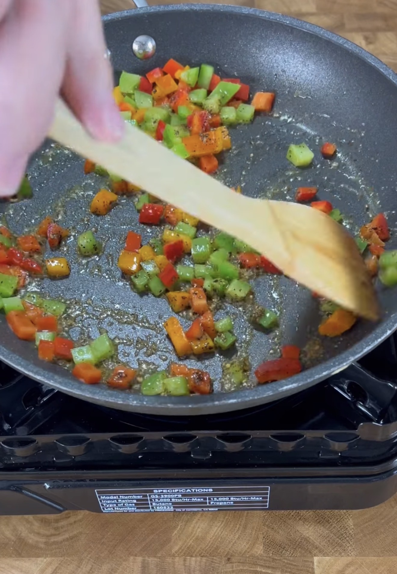 A hand stirs diced red, green, and orange bell peppers in a frying pan with a wooden spoon on a portable stove, beginning to sauté the vegetables.