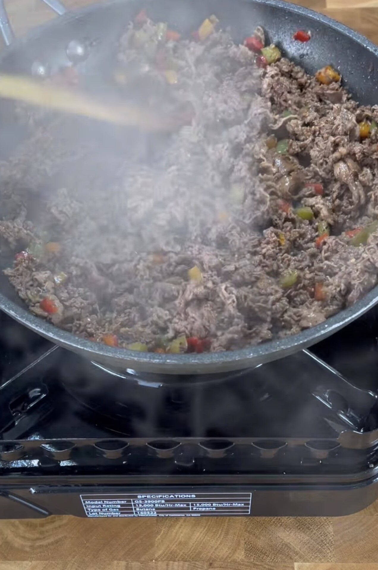 Ground beef mixed with diced vegetables cooking in a frying pan on a portable stove, with steam rising and a wooden spoon stirring the mixture.