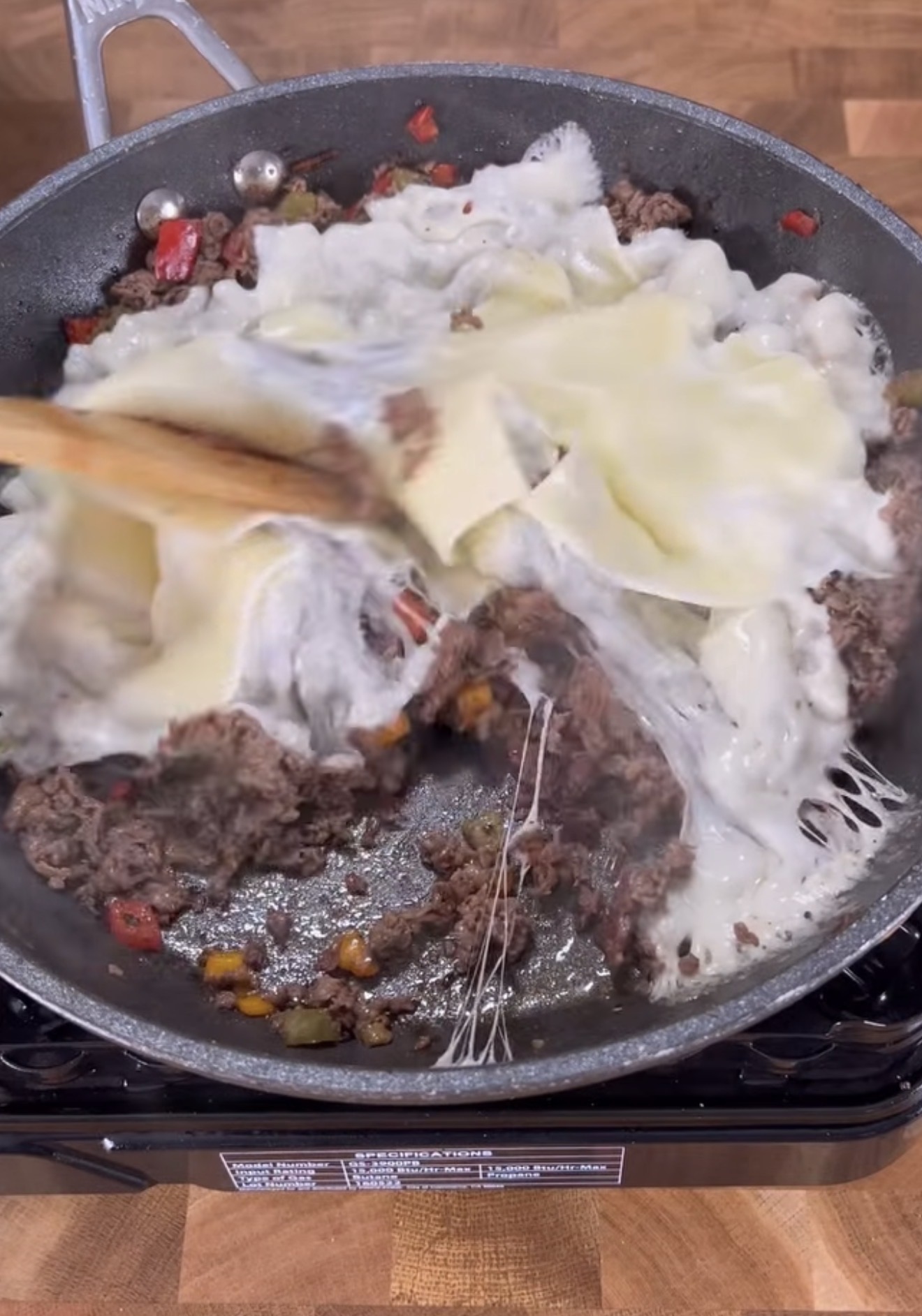 Ground beef, diced red peppers, and other vegetables cooking in a skillet, topped with melting slices of white cheese, being stirred with a wooden spoon on a wooden countertop.