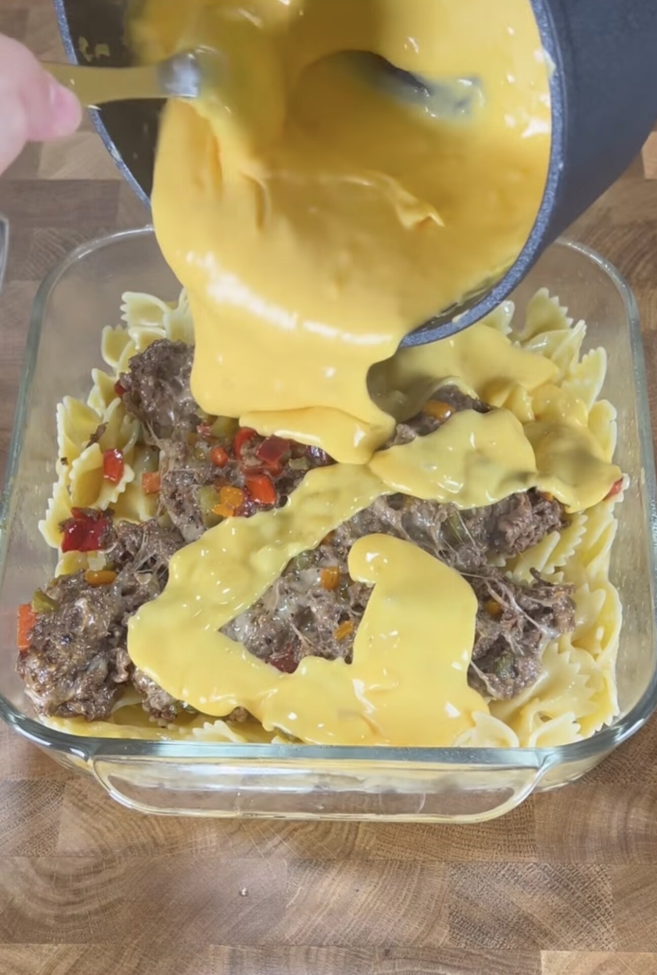 A baking dish filled with bowtie pasta, cooked ground beef, and diced bell peppers is being covered with creamy yellow cheese sauce poured from a pot.