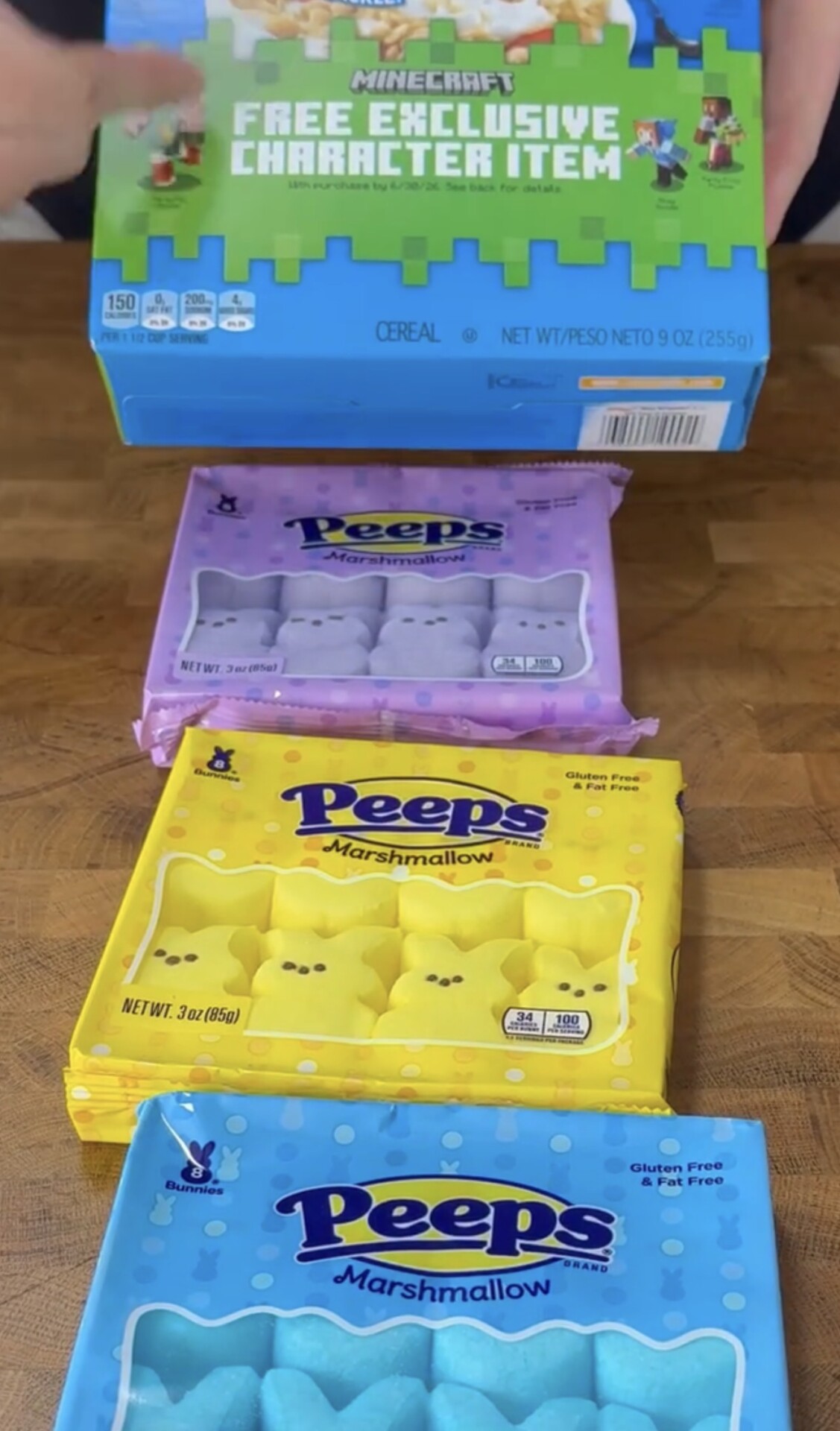 A person holds a Minecraft cereal box above three packs of Peeps marshmallow candies in purple, yellow, and blue, all placed on a wooden surface.