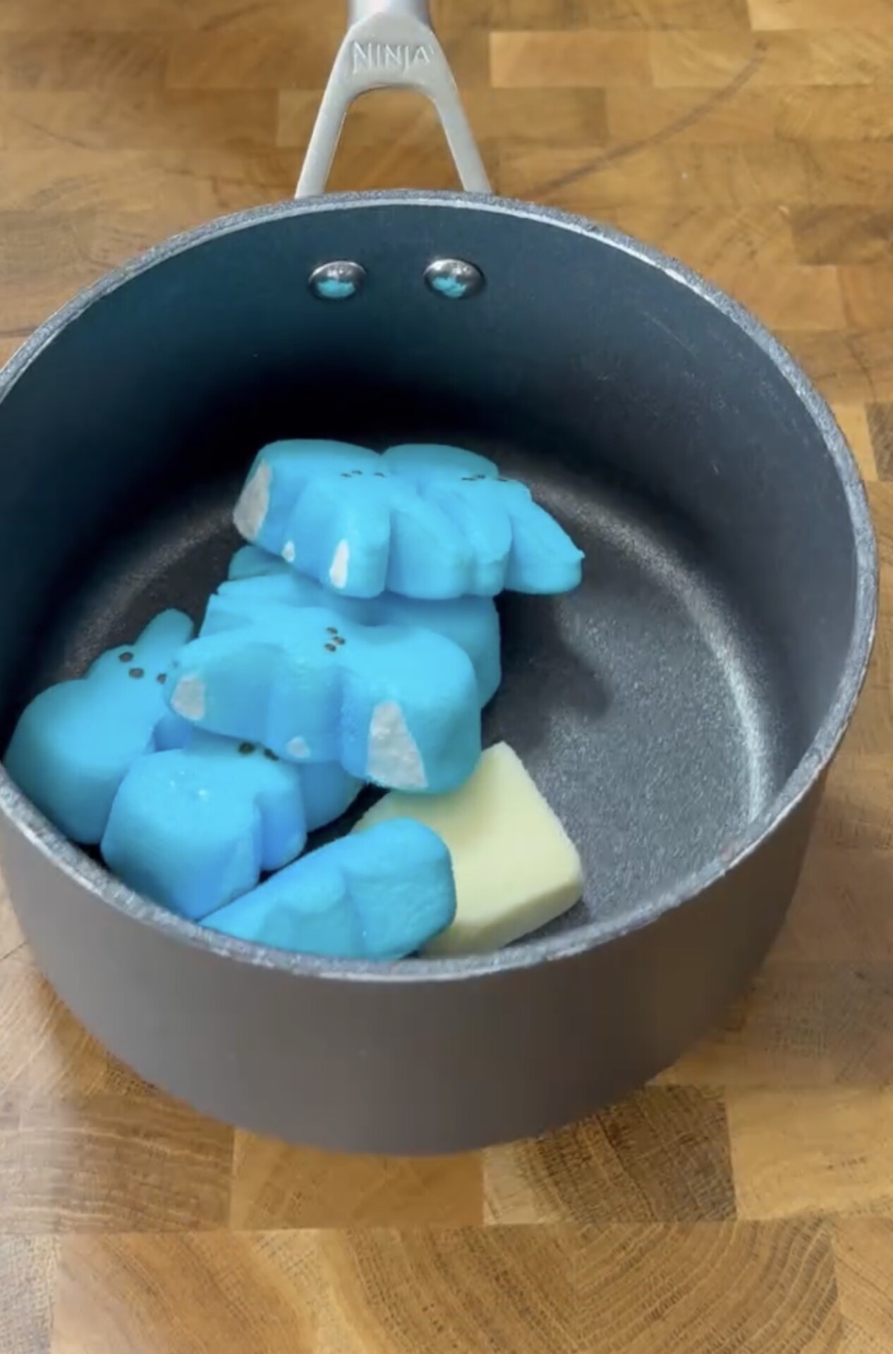 A saucepan on a wooden counter contains blue marshmallow bunny Peeps and a chunk of butter, ready to be melted together.