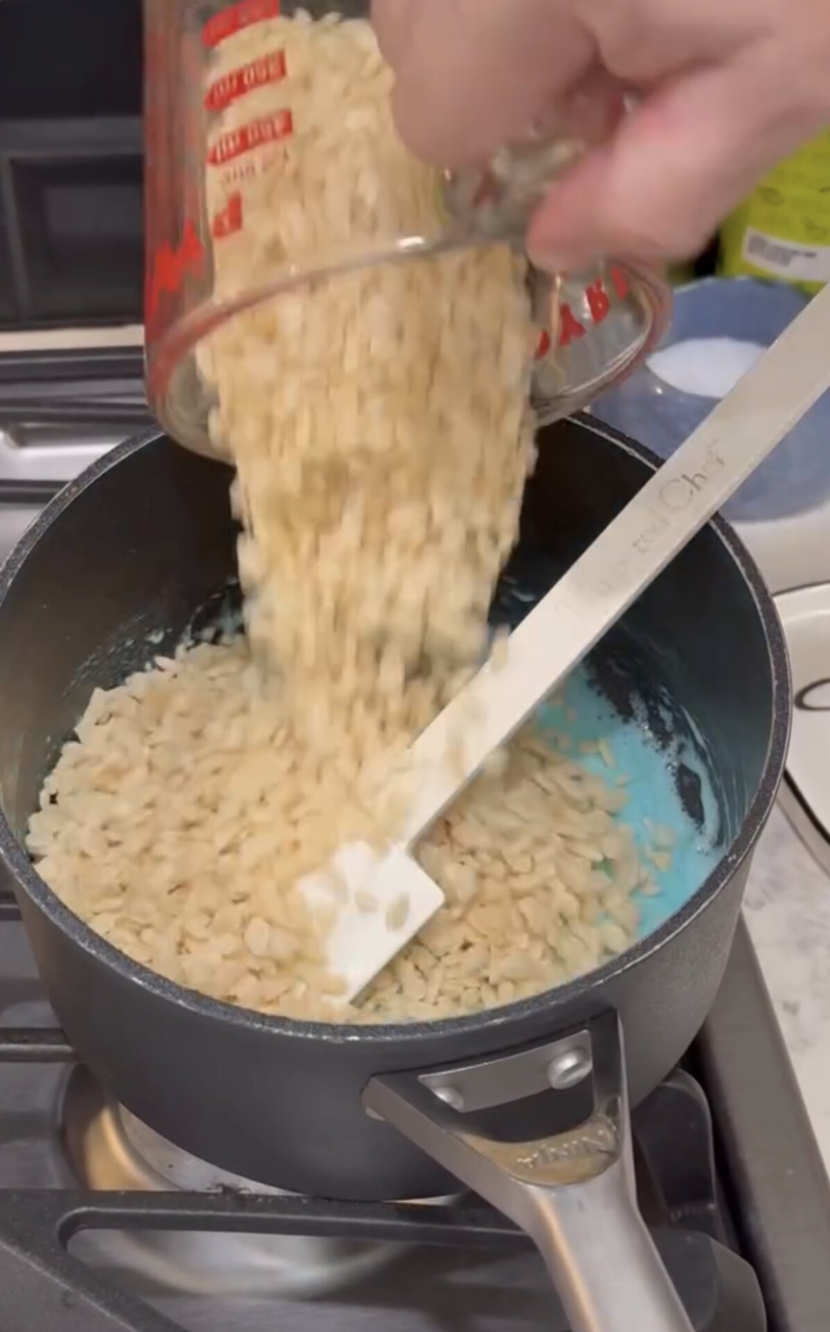 A hand pours rice cereal from a measuring cup into a saucepan with melted marshmallows, stirring with a white spatula, on a stovetop.