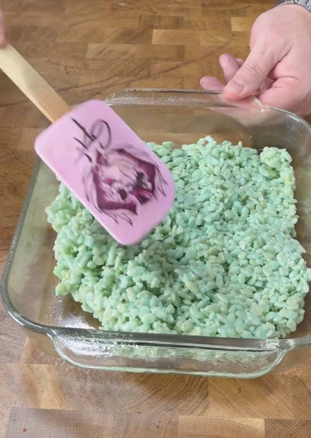 A person spreads a mixture of light green crispy rice cereal in a square glass baking dish using a pink spatula with a design on it, on a wooden surface.