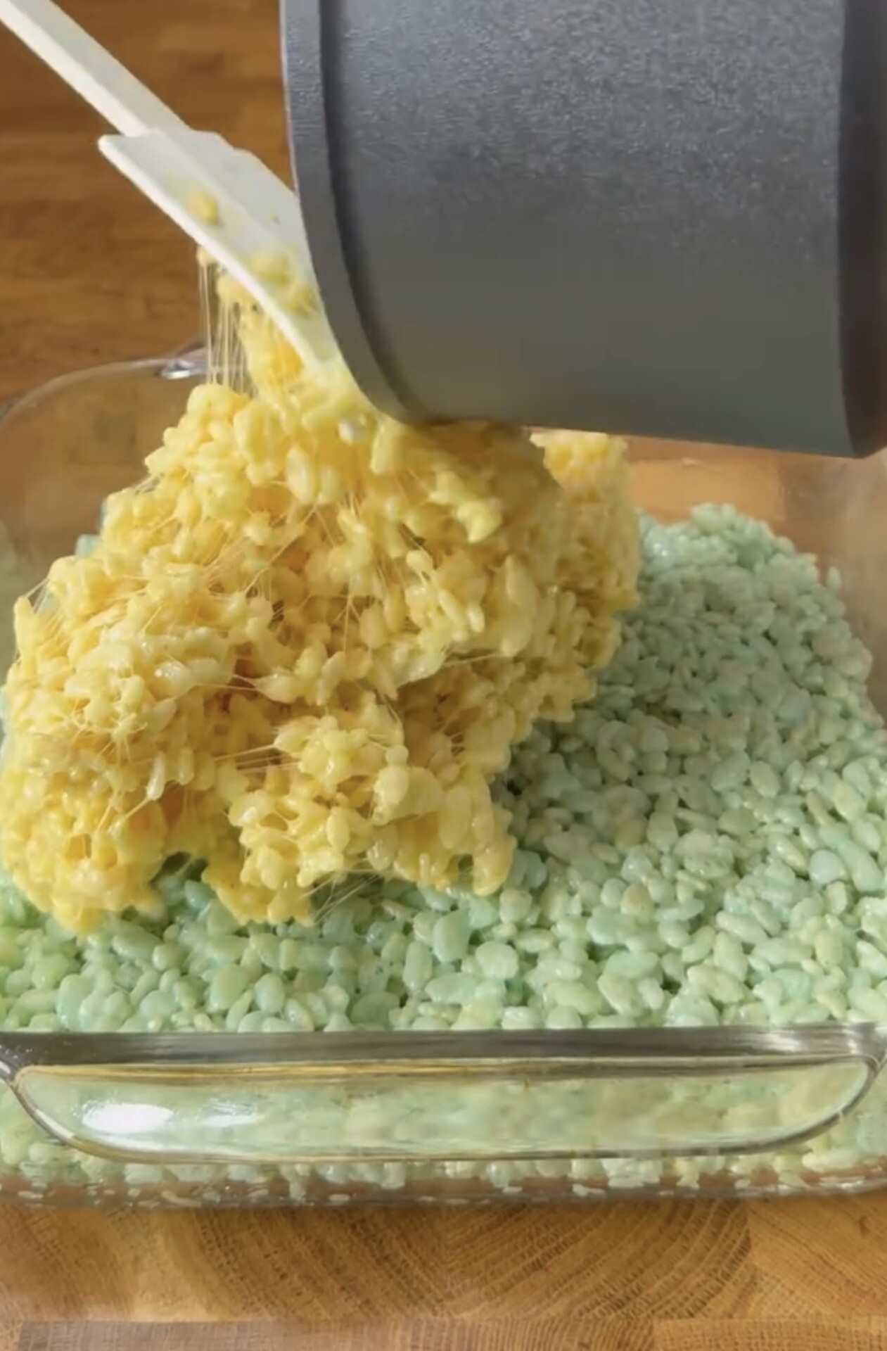 A pot of melted yellow marshmallow and cereal mixture is being poured over a layer of blue-green cereal treats in a glass baking dish on a wooden surface.