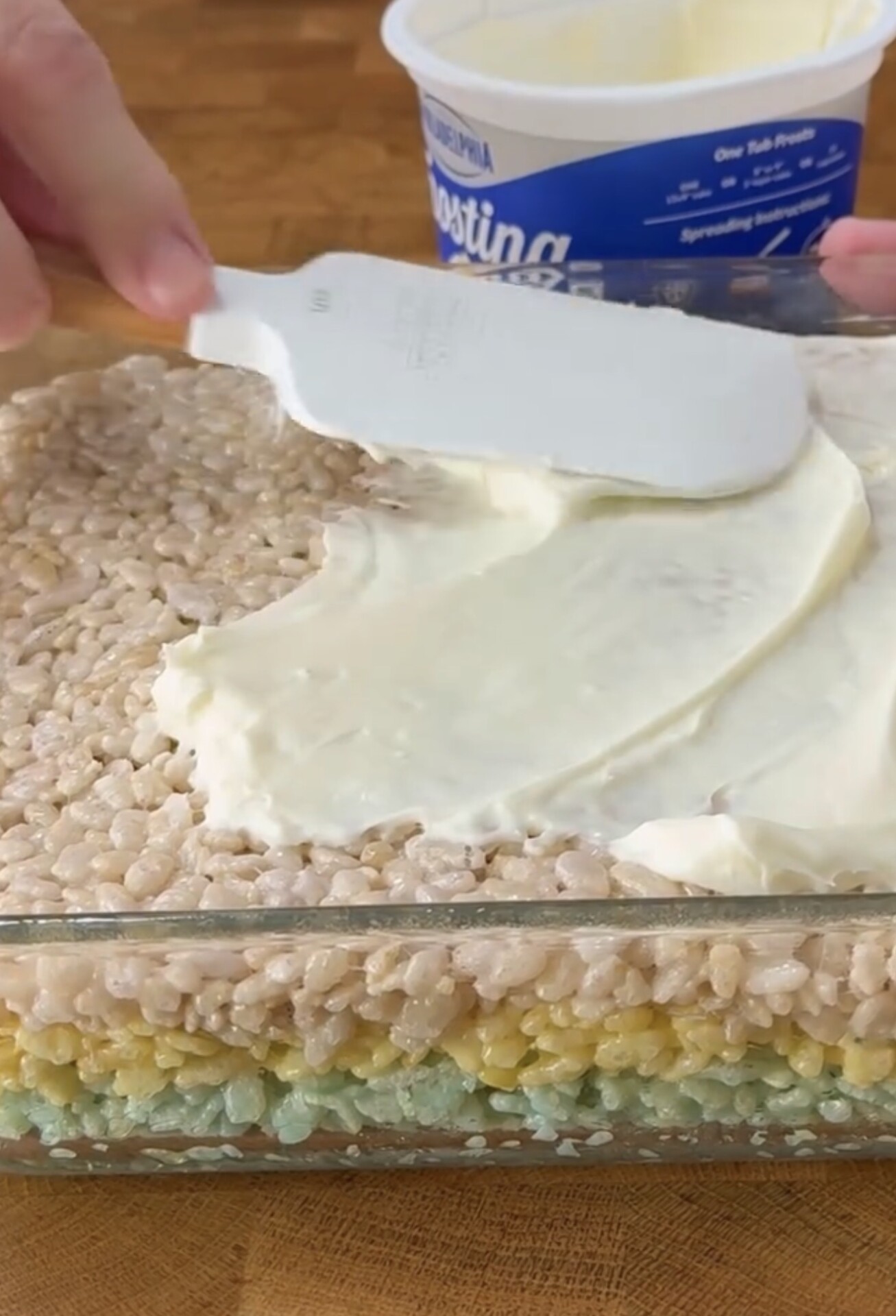 A person spreads white frosting over a layered Rice Krispies treat in a glass dish, with a container of frosting visible in the background.