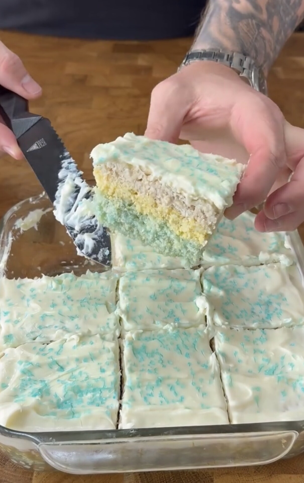 A person lifts a slice of frosted, pastel-colored layered cake from a glass baking dish. The cake is topped with white icing and blue sprinkles, and the remaining cake is cut into squares.