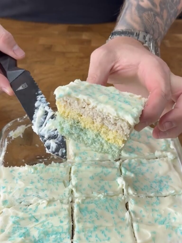 A person with tattooed arms holds a slice of layered cake with blue and yellow tiers, topped with white icing and blue sprinkles, while cutting it from a tray of similar pieces.