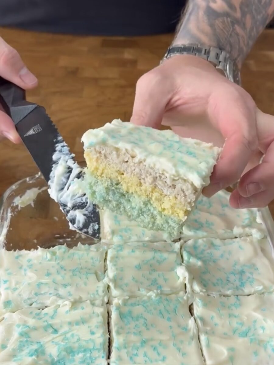 A person with tattooed arms holds a slice of layered cake with blue and yellow tiers, topped with white icing and blue sprinkles, while cutting it from a tray of similar pieces.