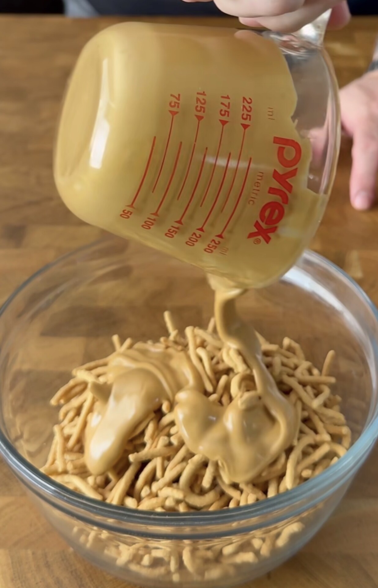 A hand pours thick, creamy peanut butter from a Pyrex measuring cup into a glass bowl filled with crunchy chow mein noodles on a wooden surface.
