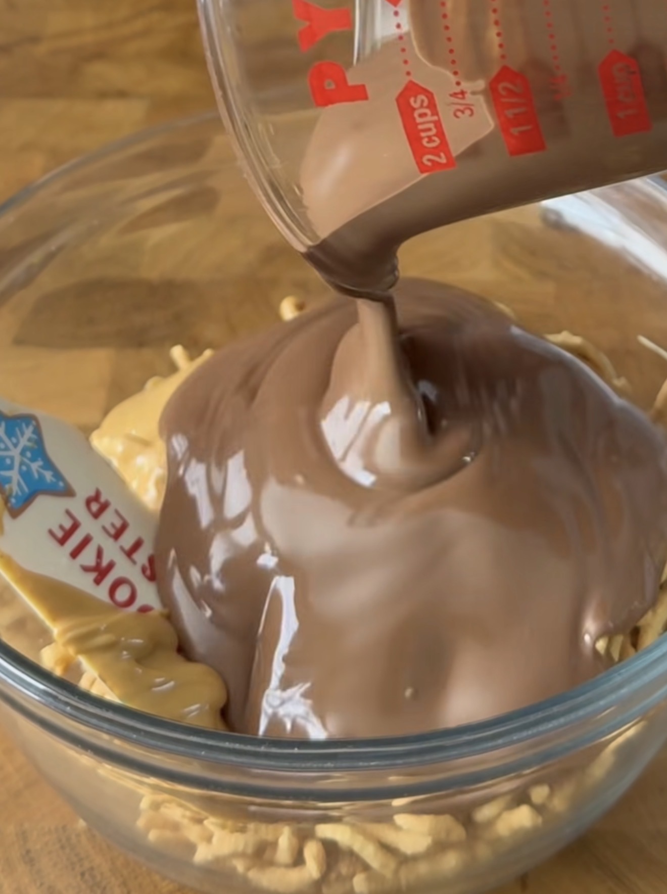 A glass measuring cup pours melted chocolate over a glass bowl containing peanut butter and crunchy noodles. A spatula labeled COOKIE MAKER rests in the bowl.