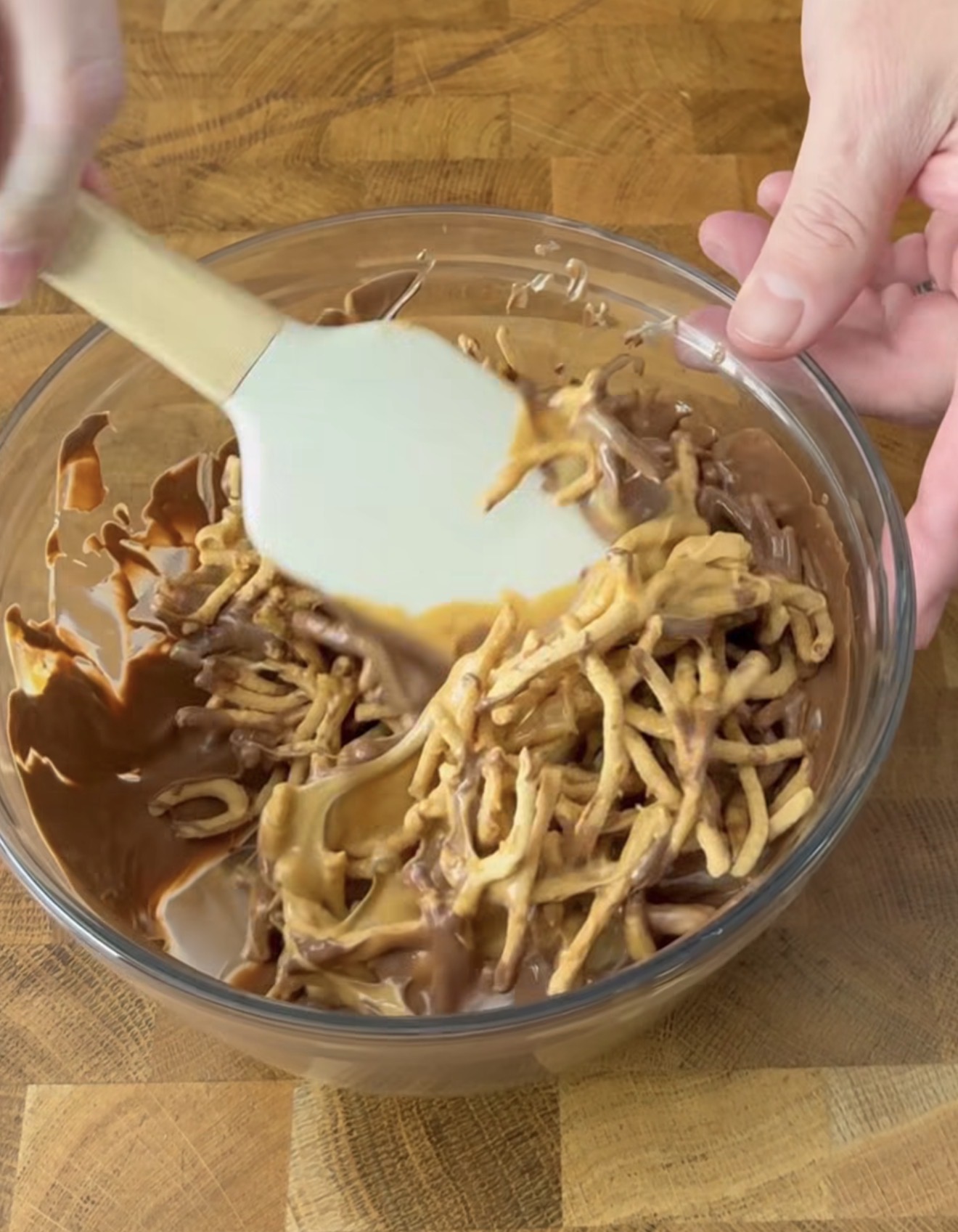 A person uses a white spatula to mix chocolate and peanut butter with crunchy chow mein noodles in a clear glass bowl on a wooden surface.