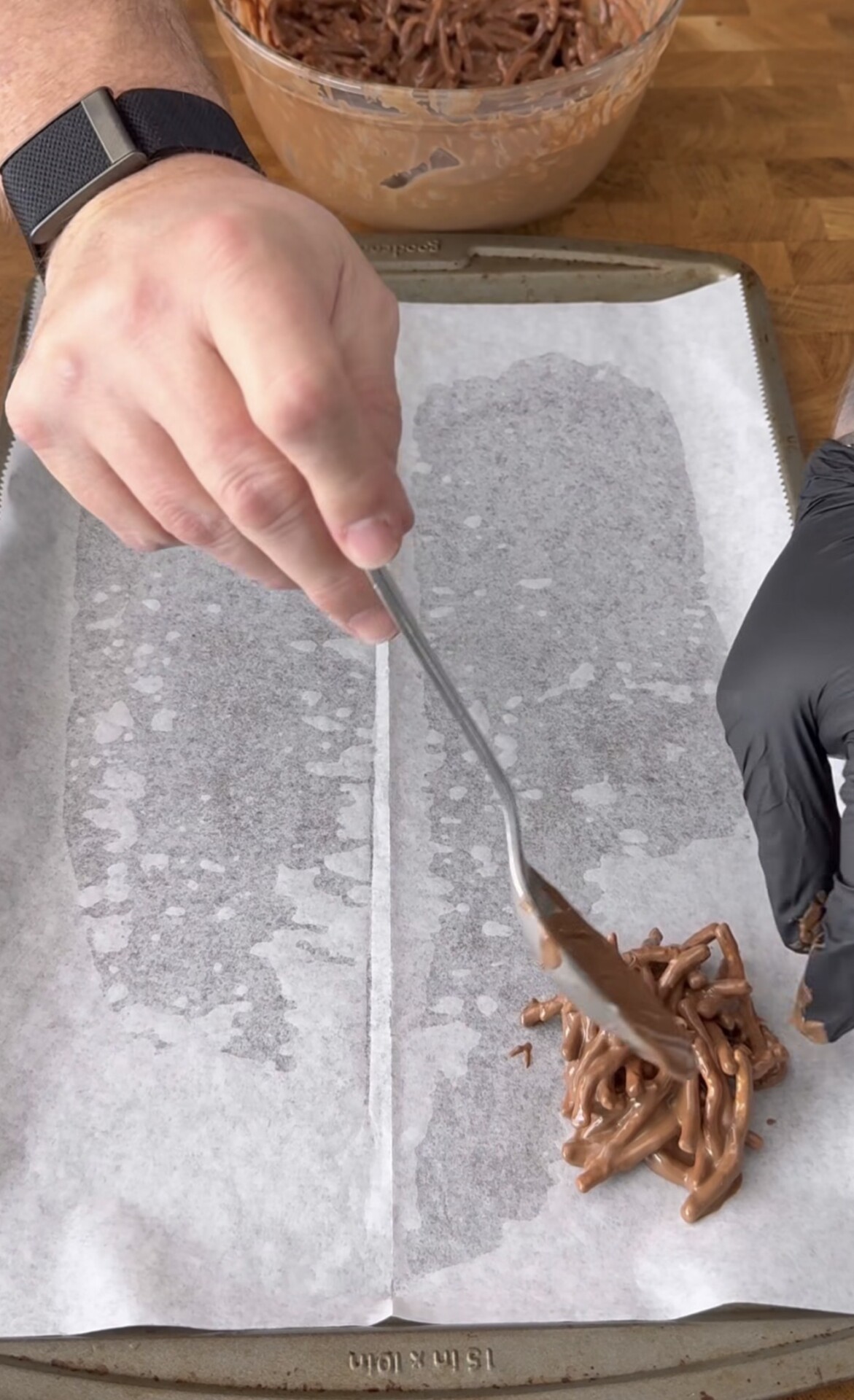 A person shapes a chocolate-coated noodle mixture onto a parchment-lined baking sheet with a spoon. One hand wears a black glove while the other holds the spoon. A bowl of the mixture is visible in the background.