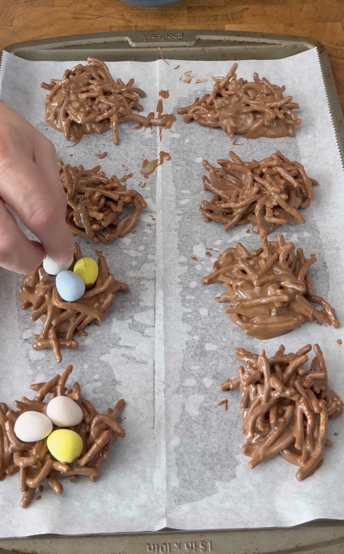 A hand places pastel candy eggs onto chocolate-covered chow mein noodle nests on a parchment-lined baking sheet, creating edible bird nests.