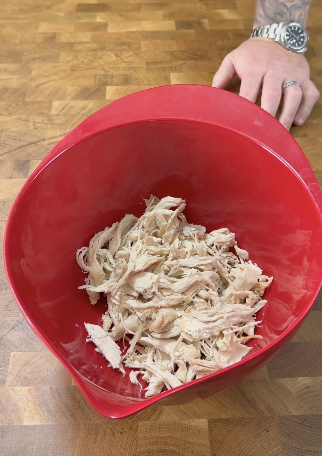 Shredded cooked chicken in a large red mixing bowl on a wooden surface, with a hand resting on the table in the background.